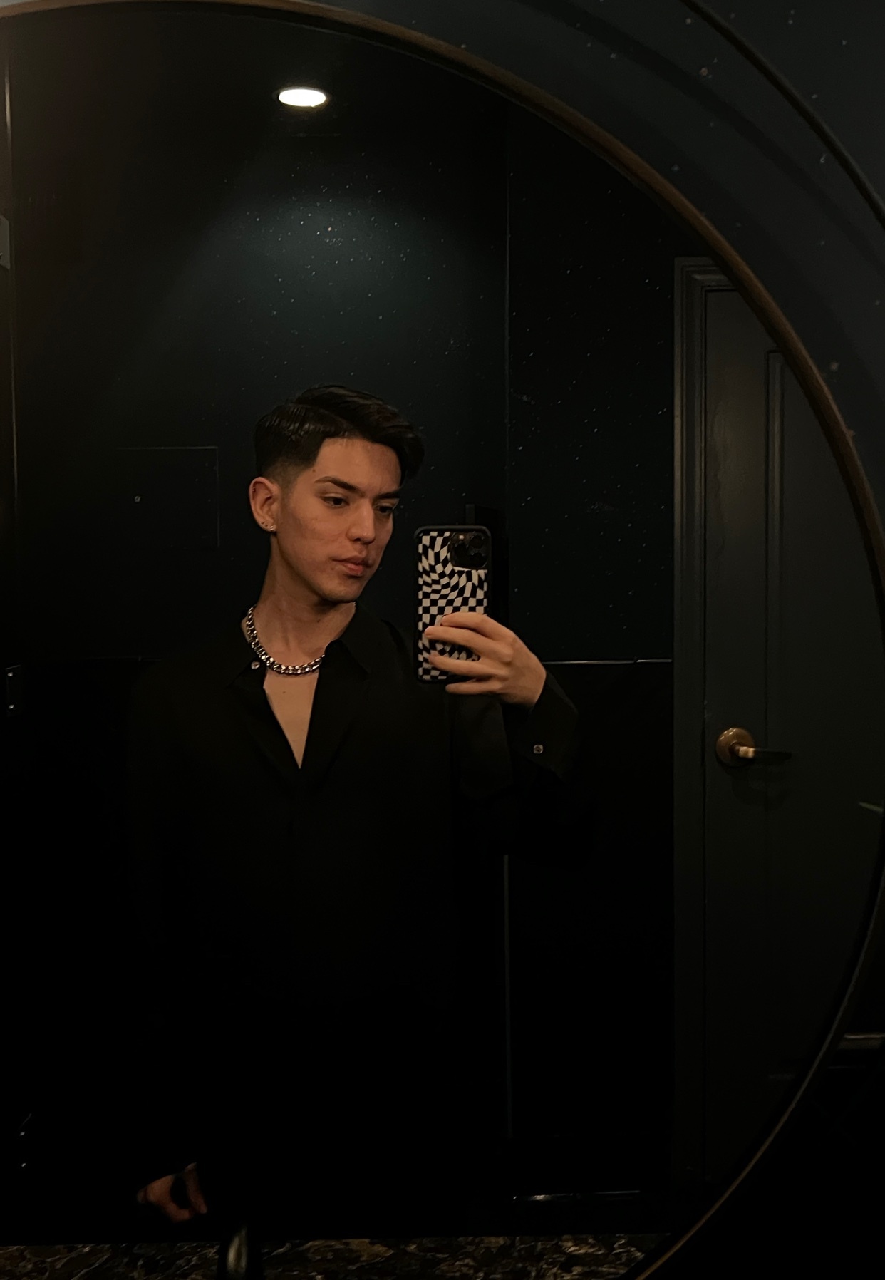 A man takes a mirror selfie in a bathroom. He is wearing a black shirt and a silver necklace.