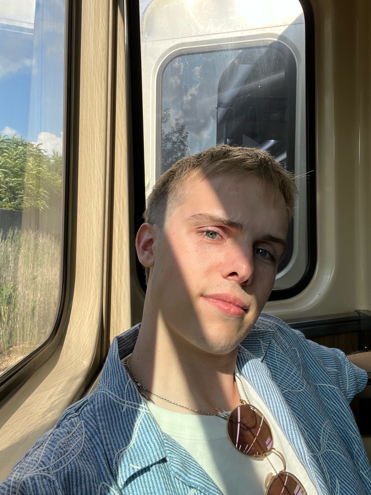 A young man with blonde hair and blue eyes takes a selfie on a train, looking wistfully at the camera. He is wearing a light blue striped shirt and sunglasses are hanging from his shirt.