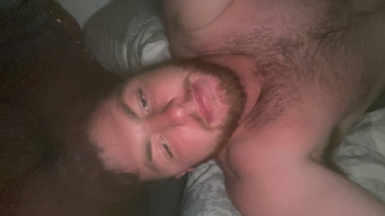 A man with a beard lies on a bed, looking up at the camera with a slight smile. His chest is bare and hairy.
