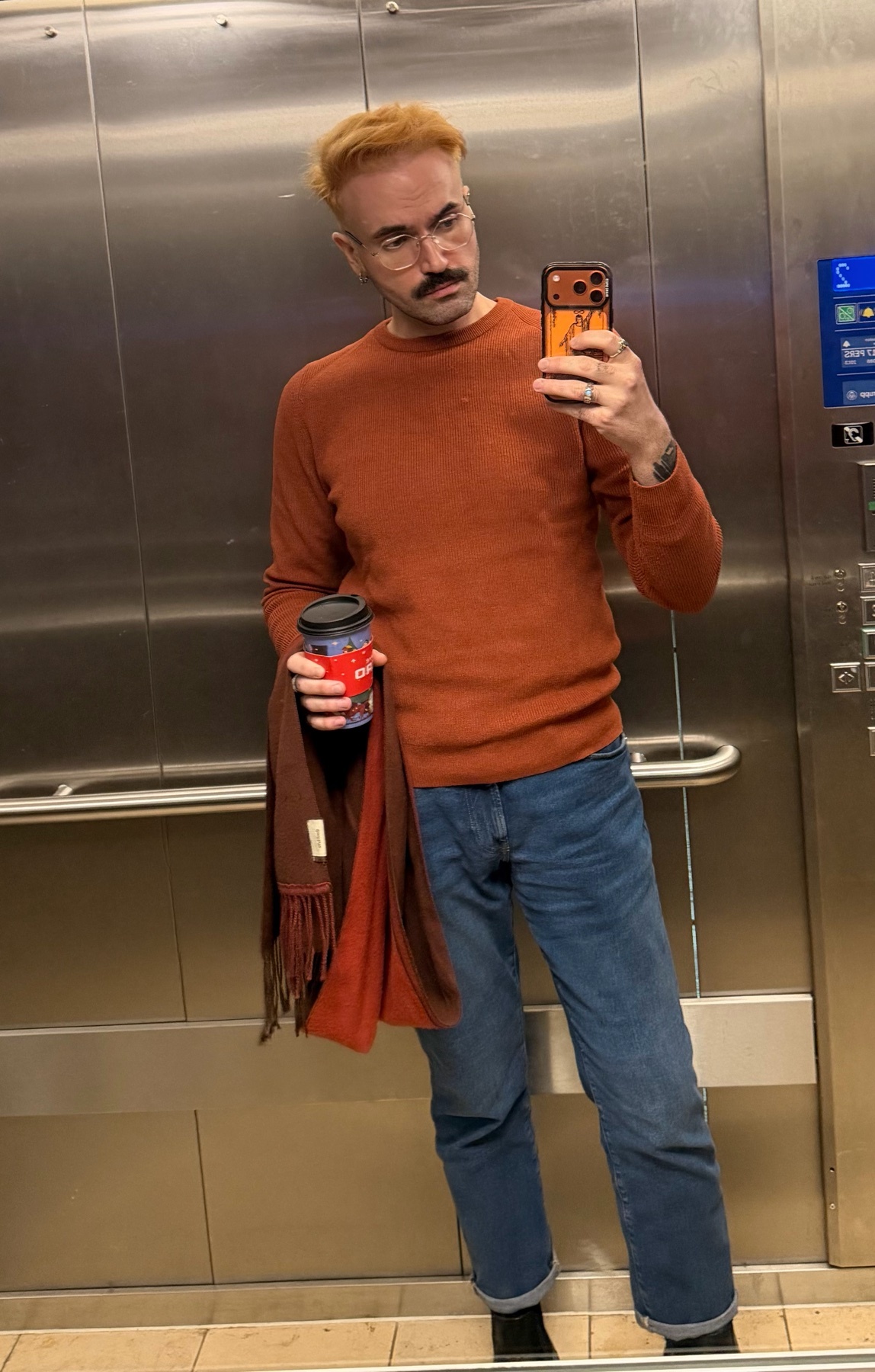 A man takes a selfie in an elevator, holding a coffee and a scarf. He is wearing a sweater and jeans.