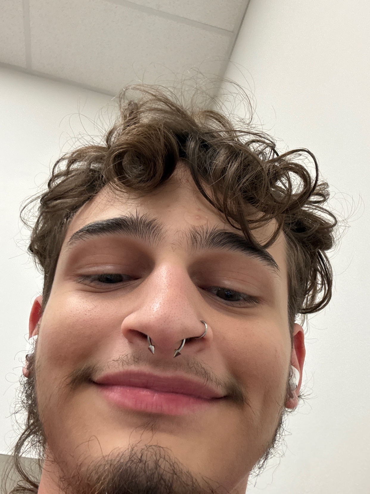A close-up selfie of a young man with curly brown hair, stubble, and a septum piercing. He is looking down and smiling slightly.
