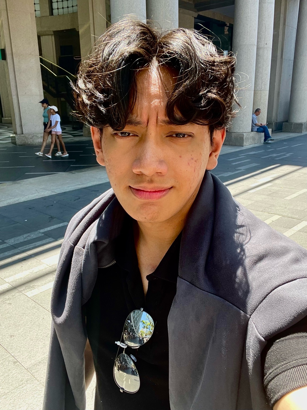 A man takes a selfie in front of a building. He is wearing a black shirt and sunglasses.