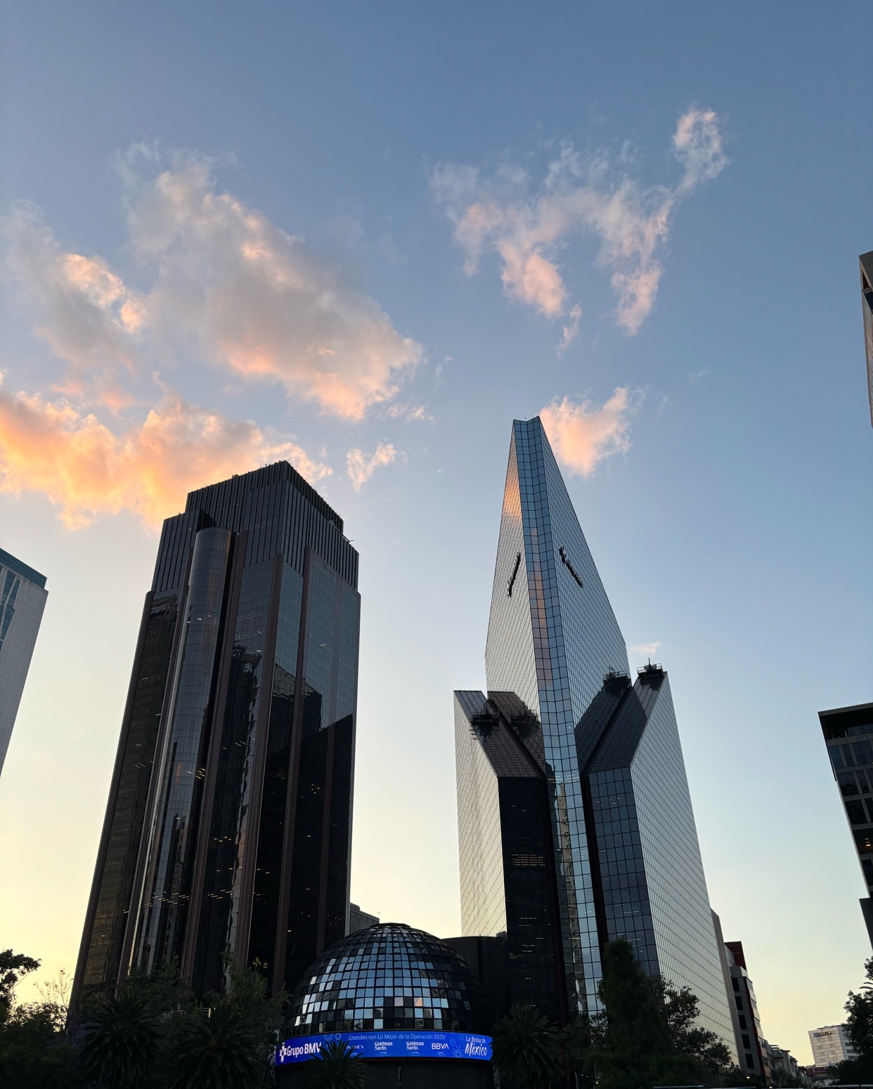A low-angle shot captures the striking modern architecture of several skyscrapers against a soft, cloud-streaked sunset sky. The buildings, with their glass facades, reflect the warm hues of the setting sun.