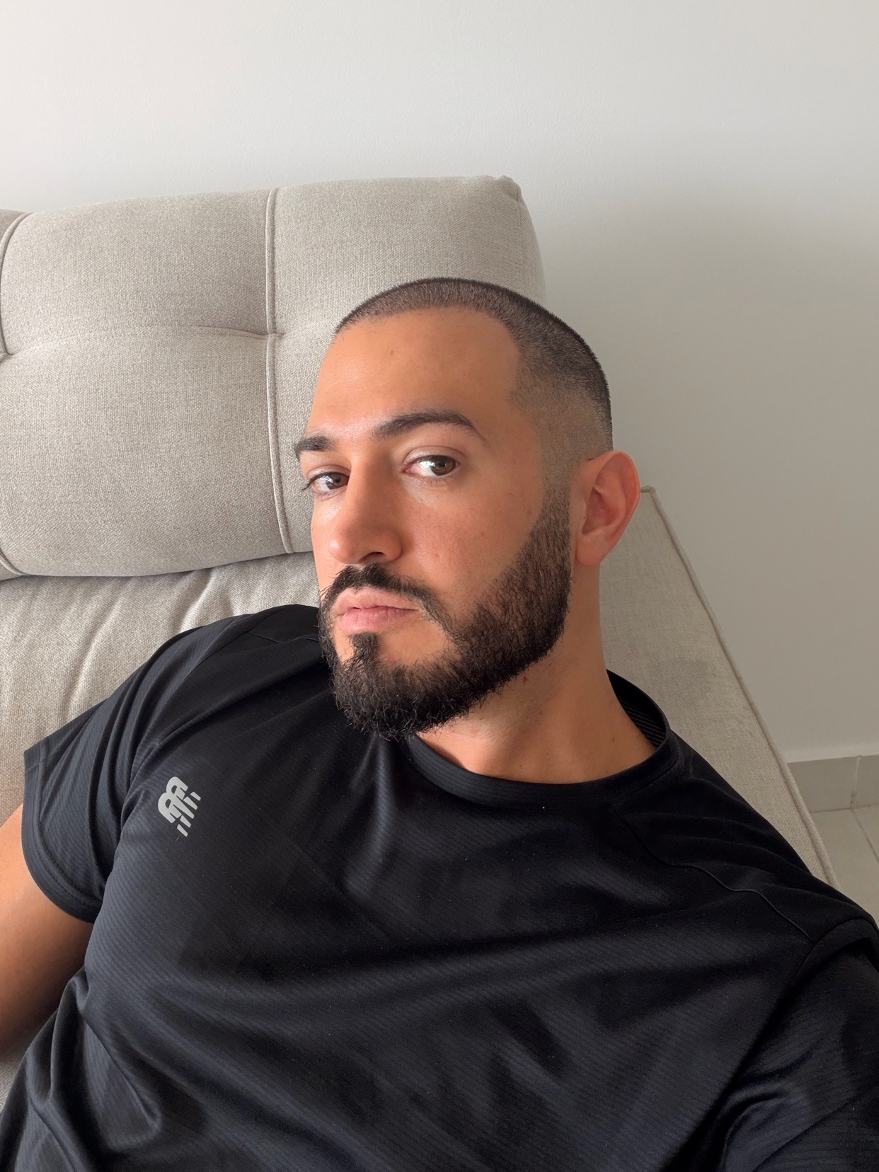 A man takes a selfie while sitting on a couch. He has a beard and is wearing a black t-shirt.
