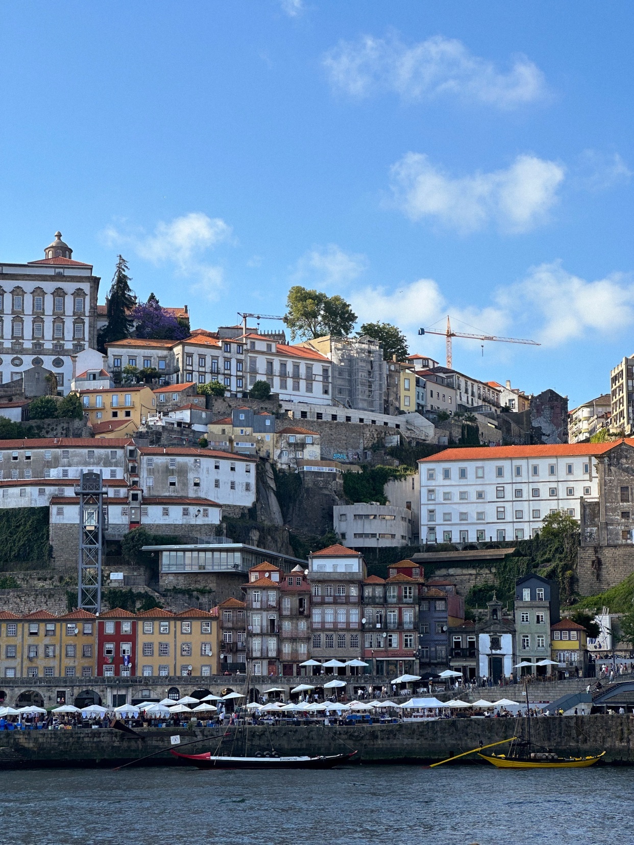 A scenic view of a European city with colorful buildings cascading down a hillside towards a river. The sky is blue with a few clouds.