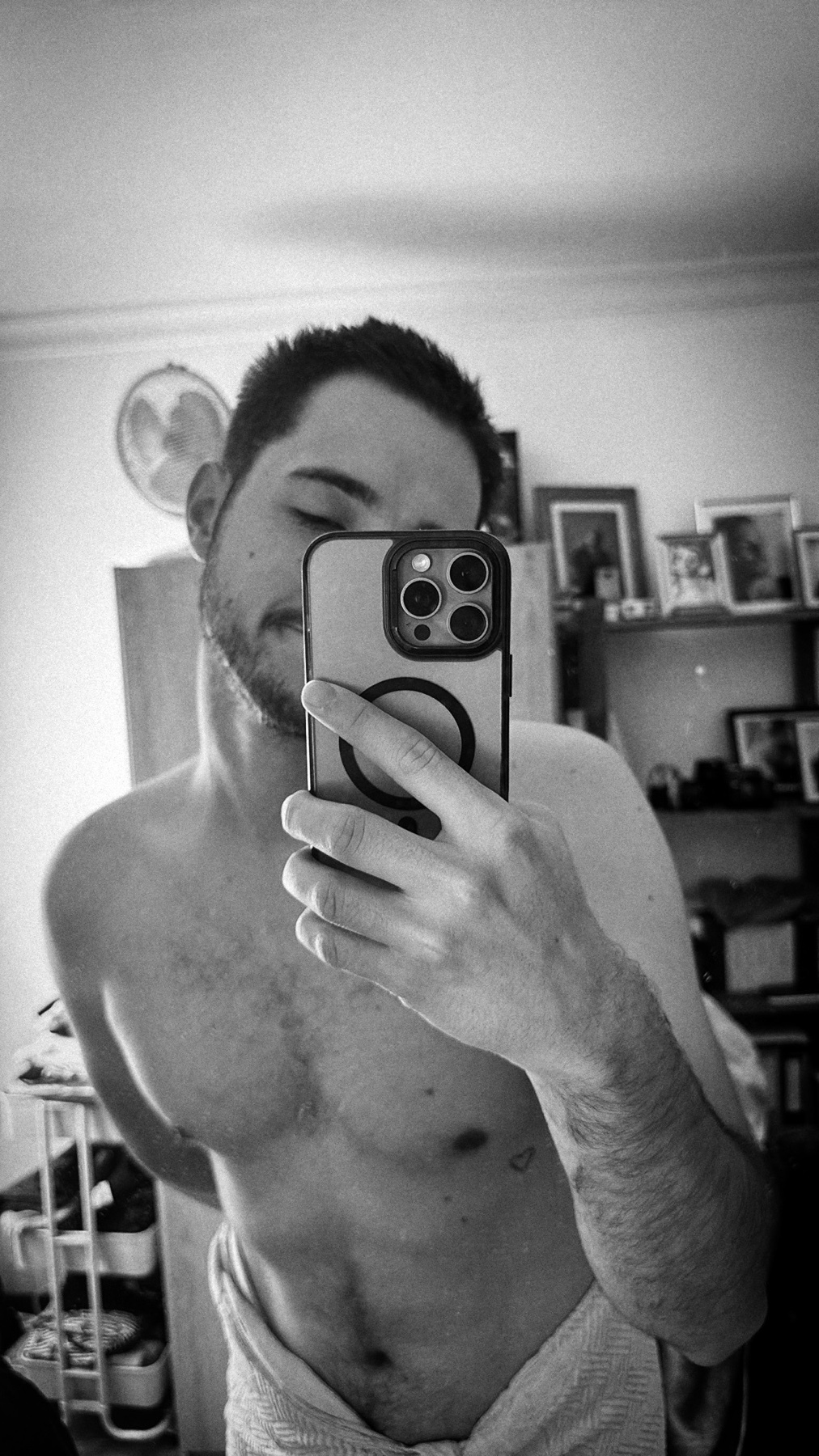 A black and white selfie of a man wearing a towel around his waist. He is holding a phone to take the picture and has stubble and a small tattoo on his chest.