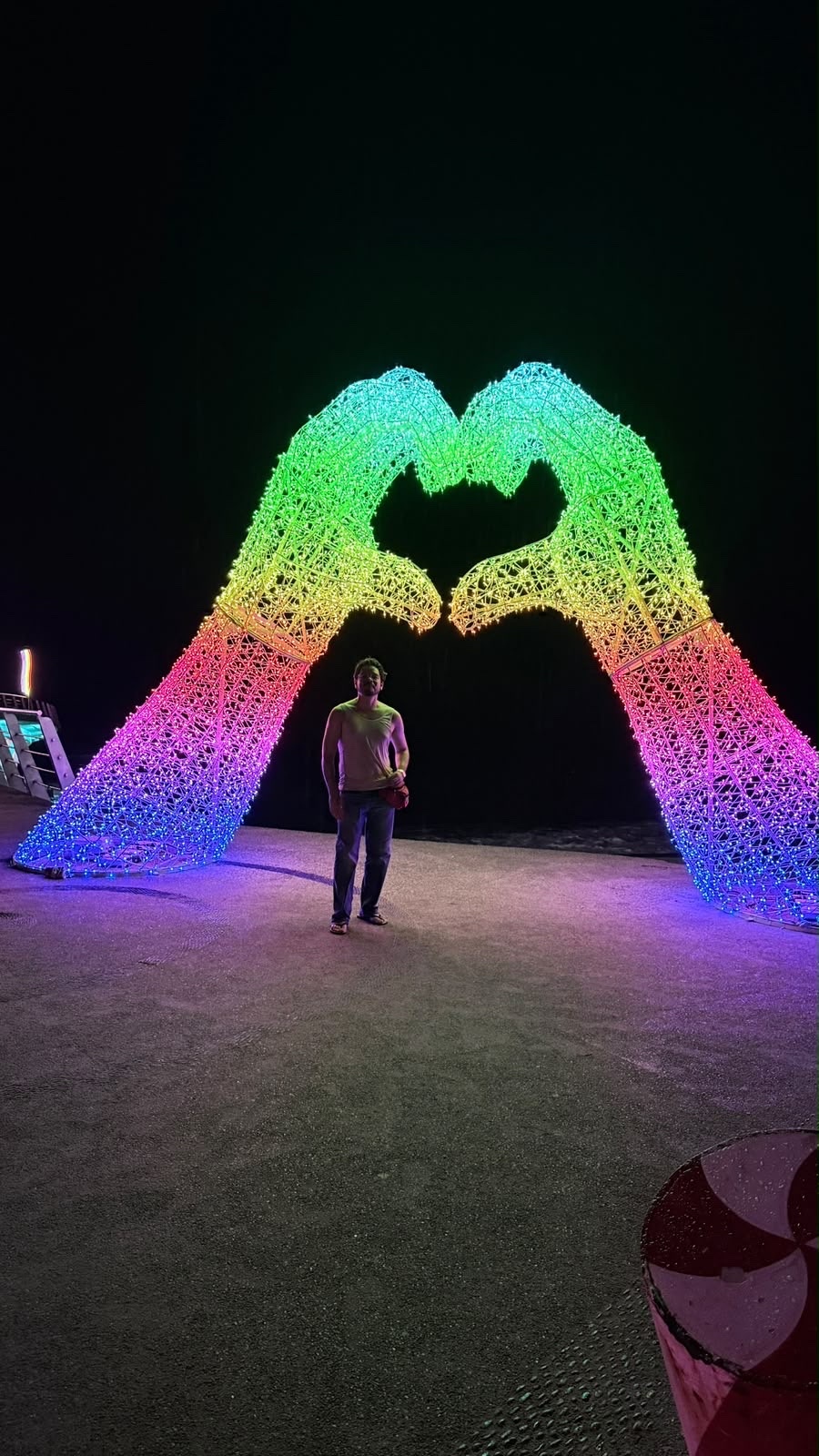 A man stands in front of a large, illuminated archway shaped like two hands forming a heart. The archway is covered in rainbow-colored lights, creating a vibrant spectacle against the dark night sky.
