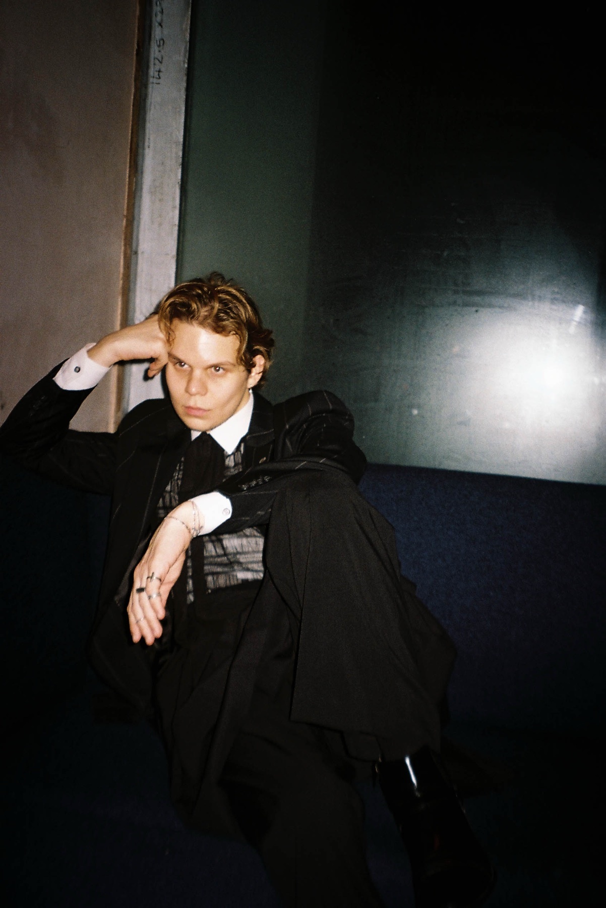 A man in a black suit and white shirt poses with his hand on his head, looking off to the side. The image has a dark, moody aesthetic.