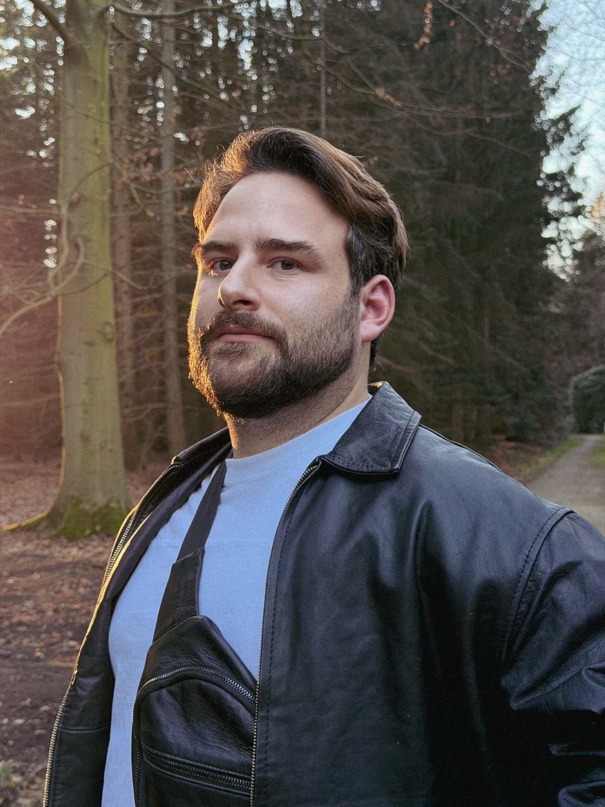 A man with a beard and mustache poses for a portrait outdoors, wearing a leather jacket over a light blue t-shirt. The background features trees and a path, suggesting a park or wooded area.
