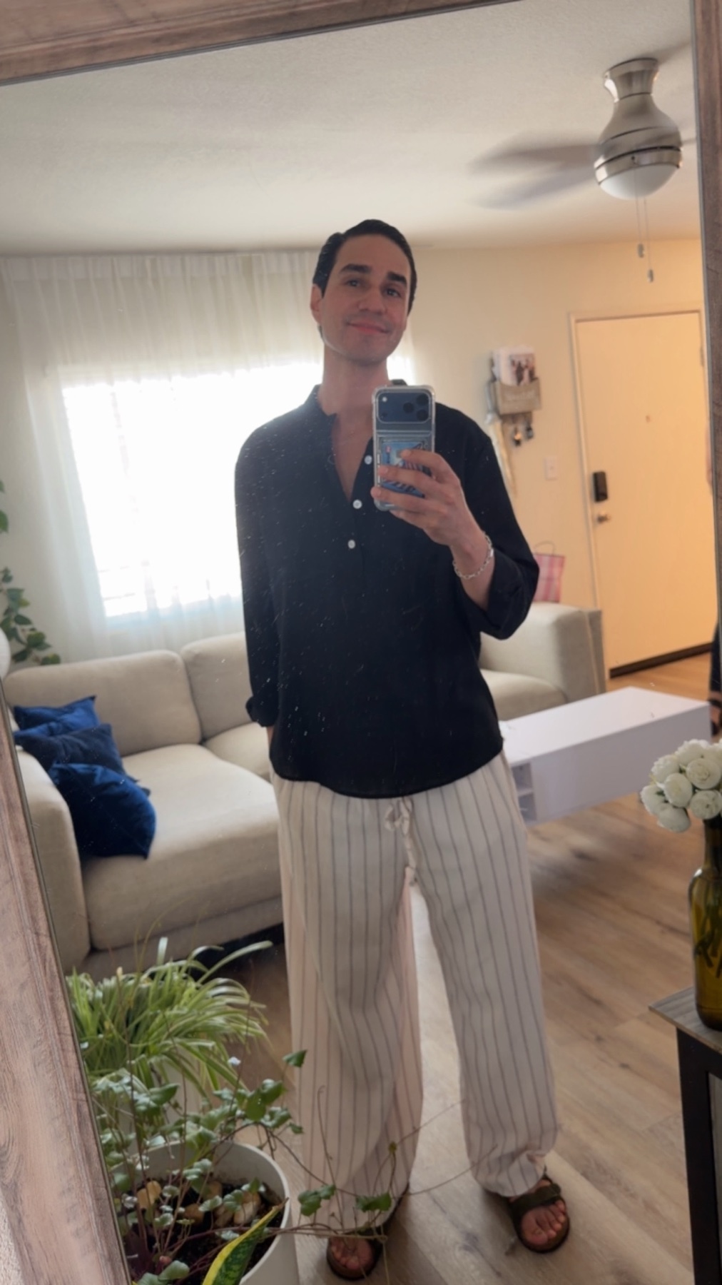 A man takes a mirror selfie in a casual outfit. He is wearing a black button-up shirt and striped pants.