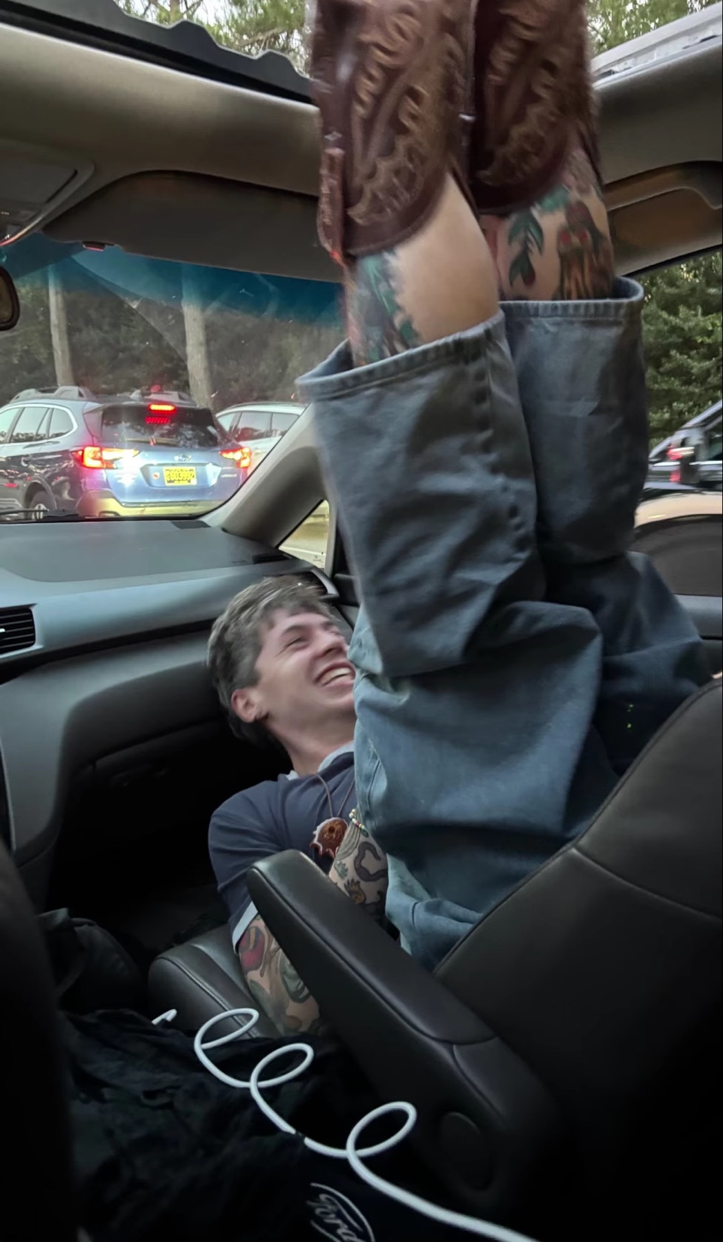 A man is laughing inside a car while another person's legs, wearing jeans and boots, are positioned above him.