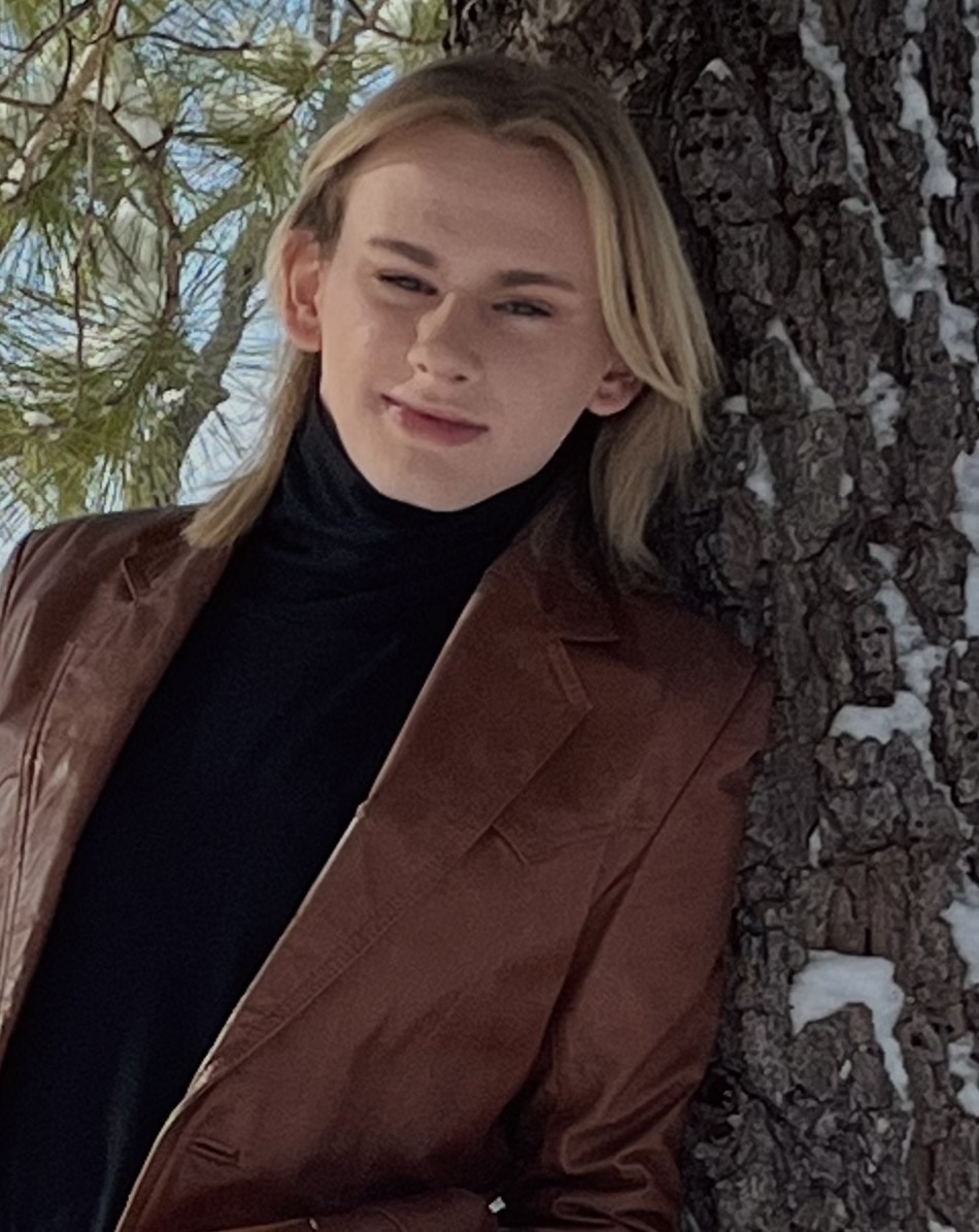 A woman poses in a brown leather jacket and black turtleneck, leaning against a snow-covered tree. The photo is taken in daylight.