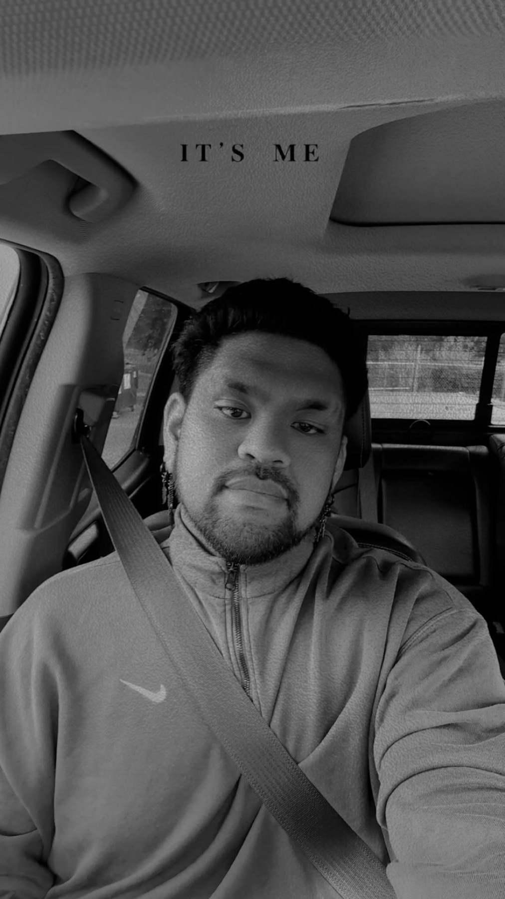 A black and white selfie of a man sitting in the driver's seat of a car. The text "IT'S ME" is overlaid at the top of the image.