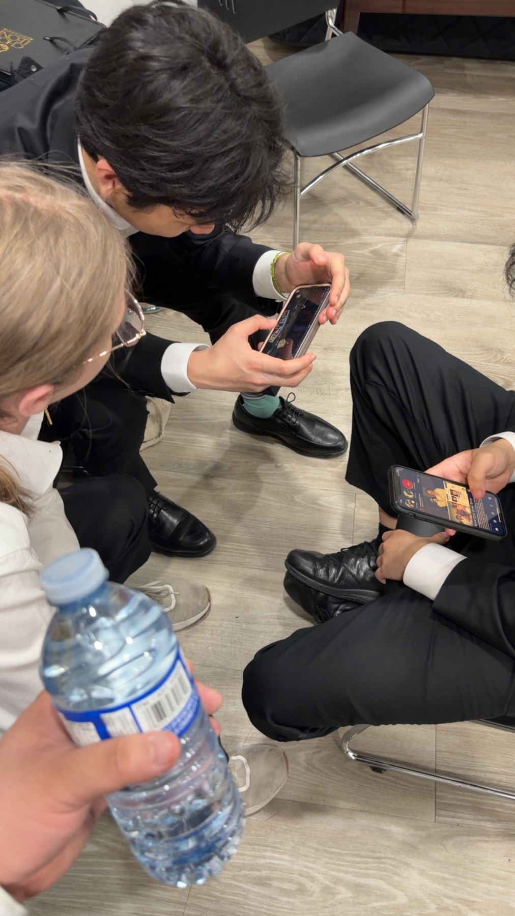 A group of friends in suits are looking at their phones, appearing exhausted after a concert. One person holds a water bottle.