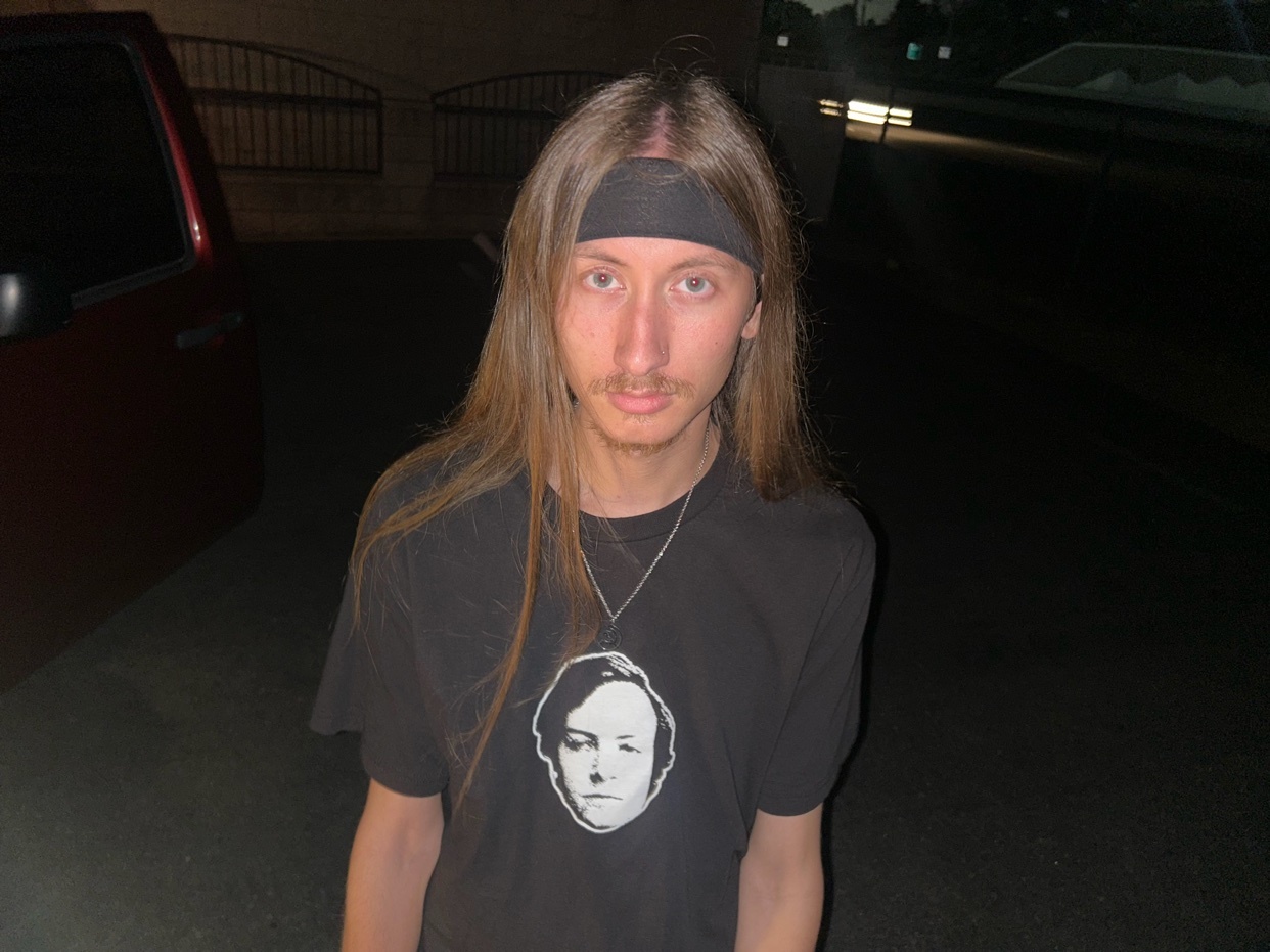 A man with long hair and a bandana poses for a photo. He is wearing a black t-shirt.