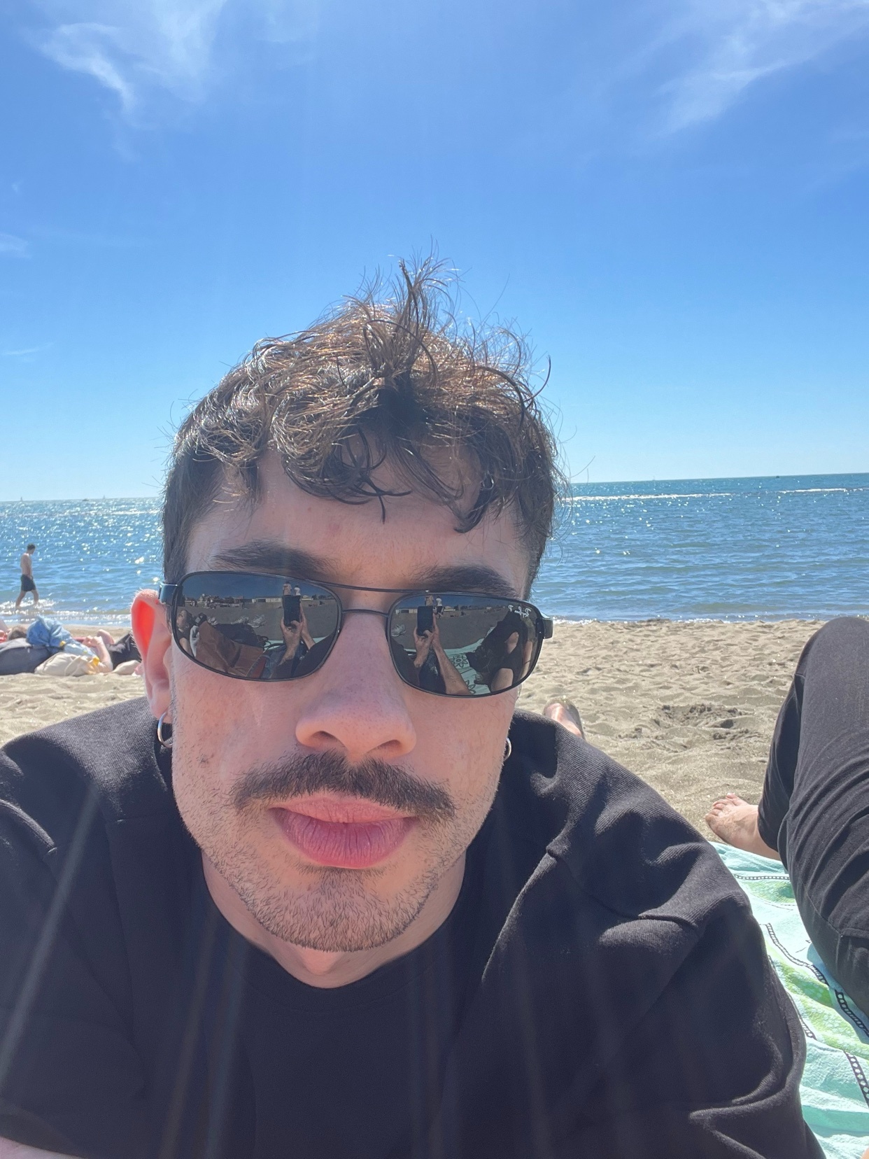 A man with a mustache and sunglasses takes a selfie on a sunny beach day. The ocean and sand are visible in the background.