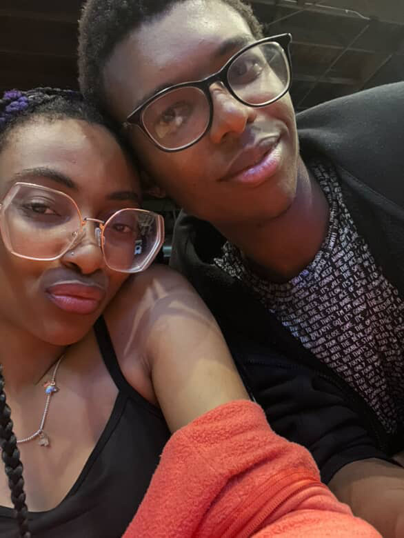 A close-up selfie shows a Black couple, with the woman in the foreground wearing large glasses and a black tank top, and the man behind her wearing glasses and a black zip-up hoodie. They are posing for the camera.