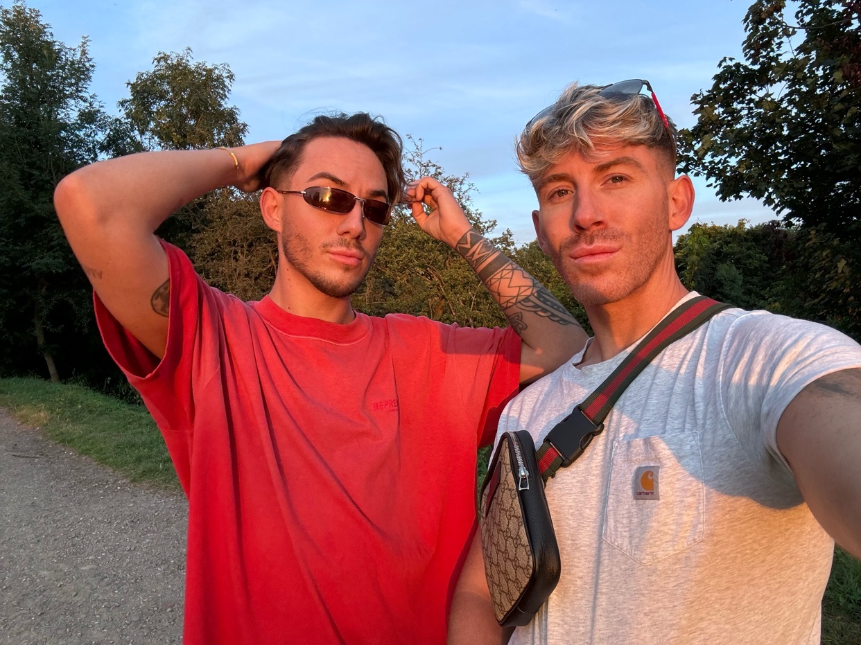 A selfie of two men posing outdoors. They are wearing sunglasses and casual clothing.