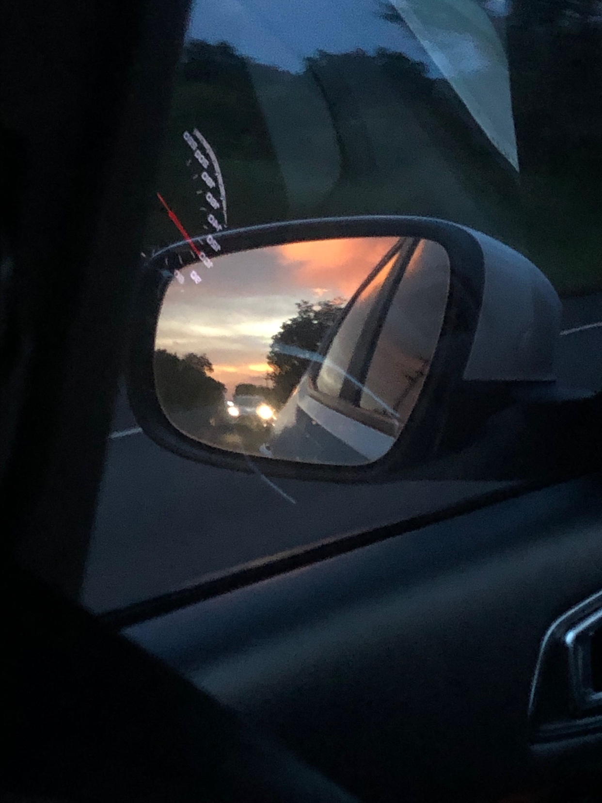 A car's side mirror reflects a vibrant sunset with colorful clouds and trees. The image captures the fleeting beauty of the sky as seen from the perspective of a moving vehicle.