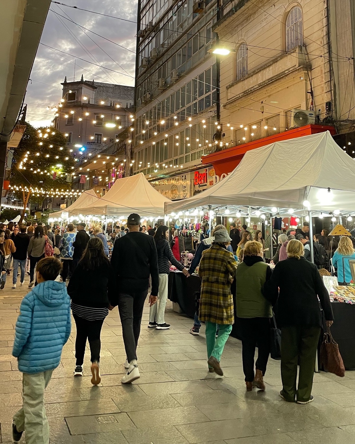 A bustling street market at night, filled with people and illuminated by string lights. The scene captures the energy of a city's evening activities.