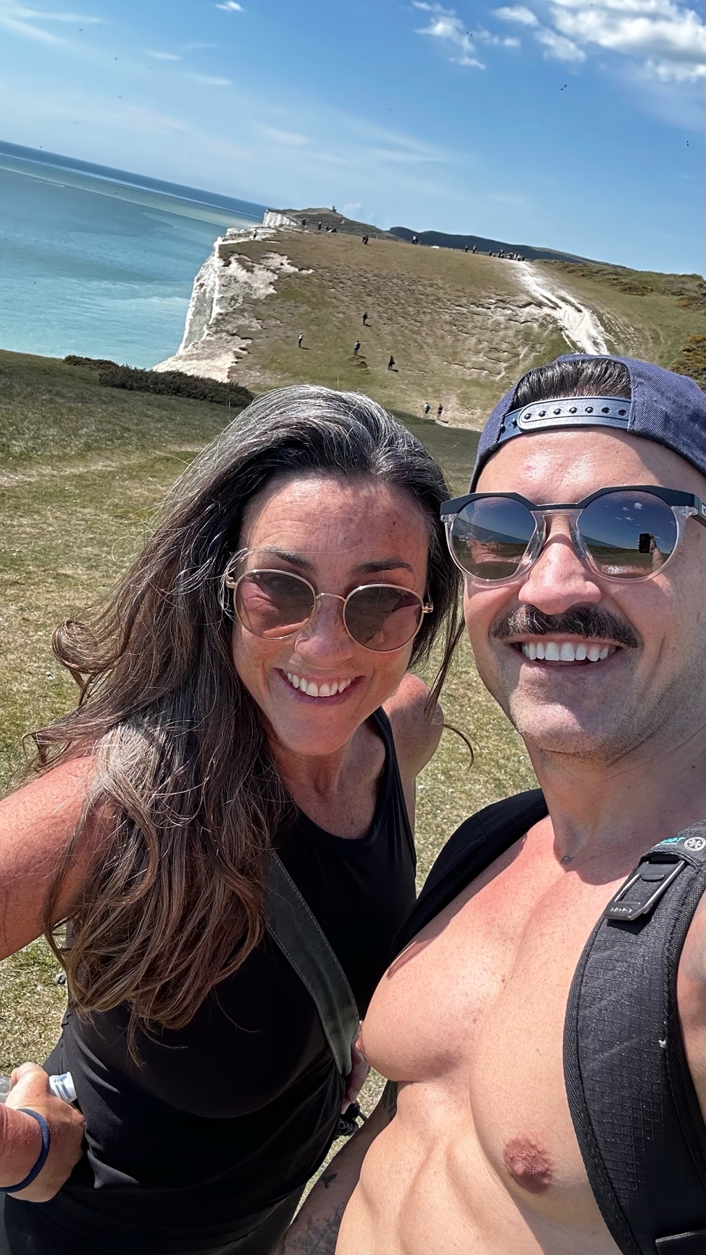 A man and woman take a selfie on a cliff overlooking the ocean on a sunny day. The man is shirtless and wearing a baseball cap and sunglasses, while the woman has long, graying hair and is also wearing sunglasses.