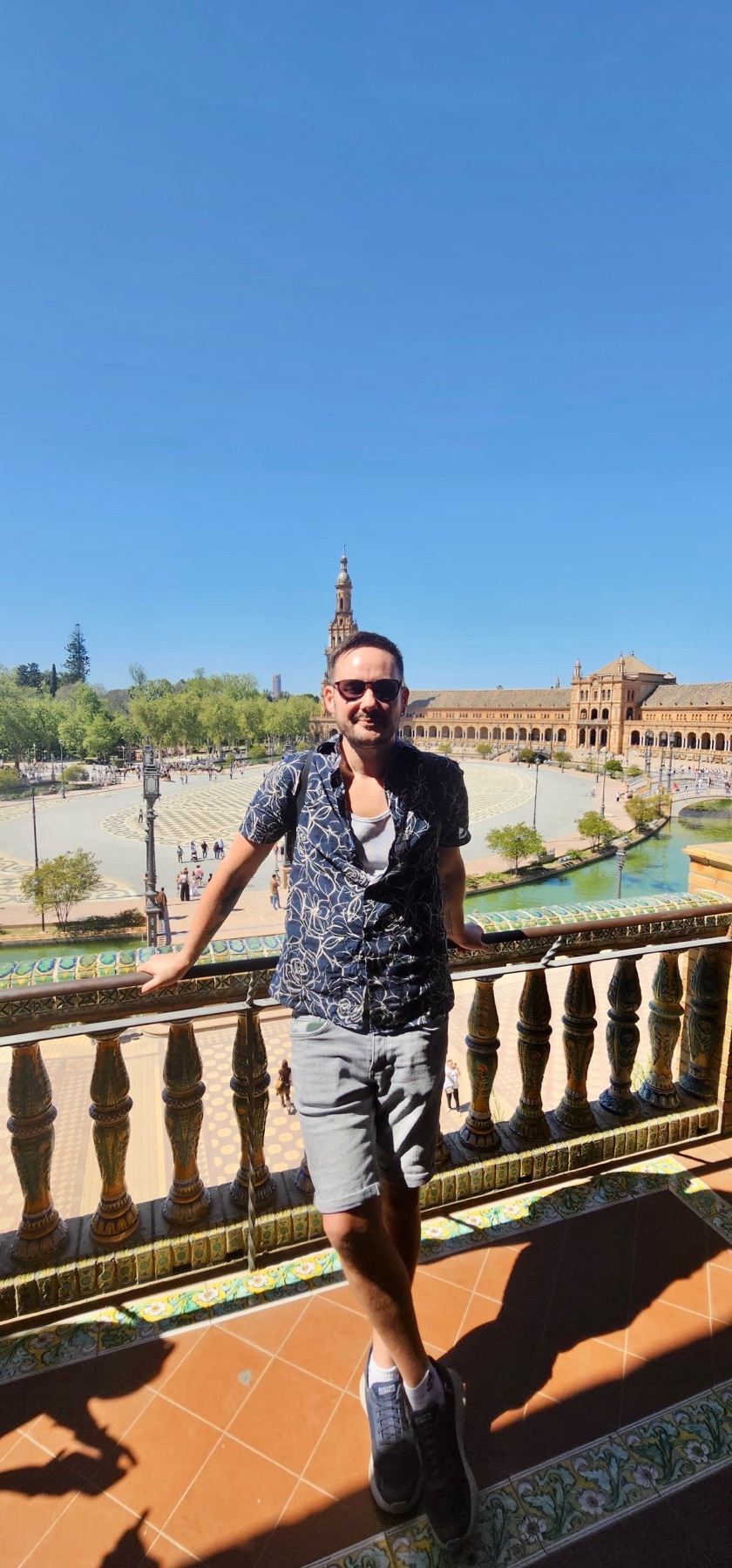 A man poses for a photo on a sunny day in front of a large, ornate building with a plaza and water features.