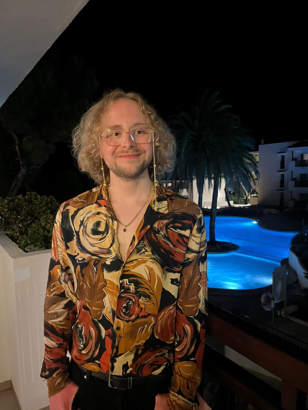 A man with curly hair and glasses poses in a floral shirt at night. The photo was taken in Mallorca.