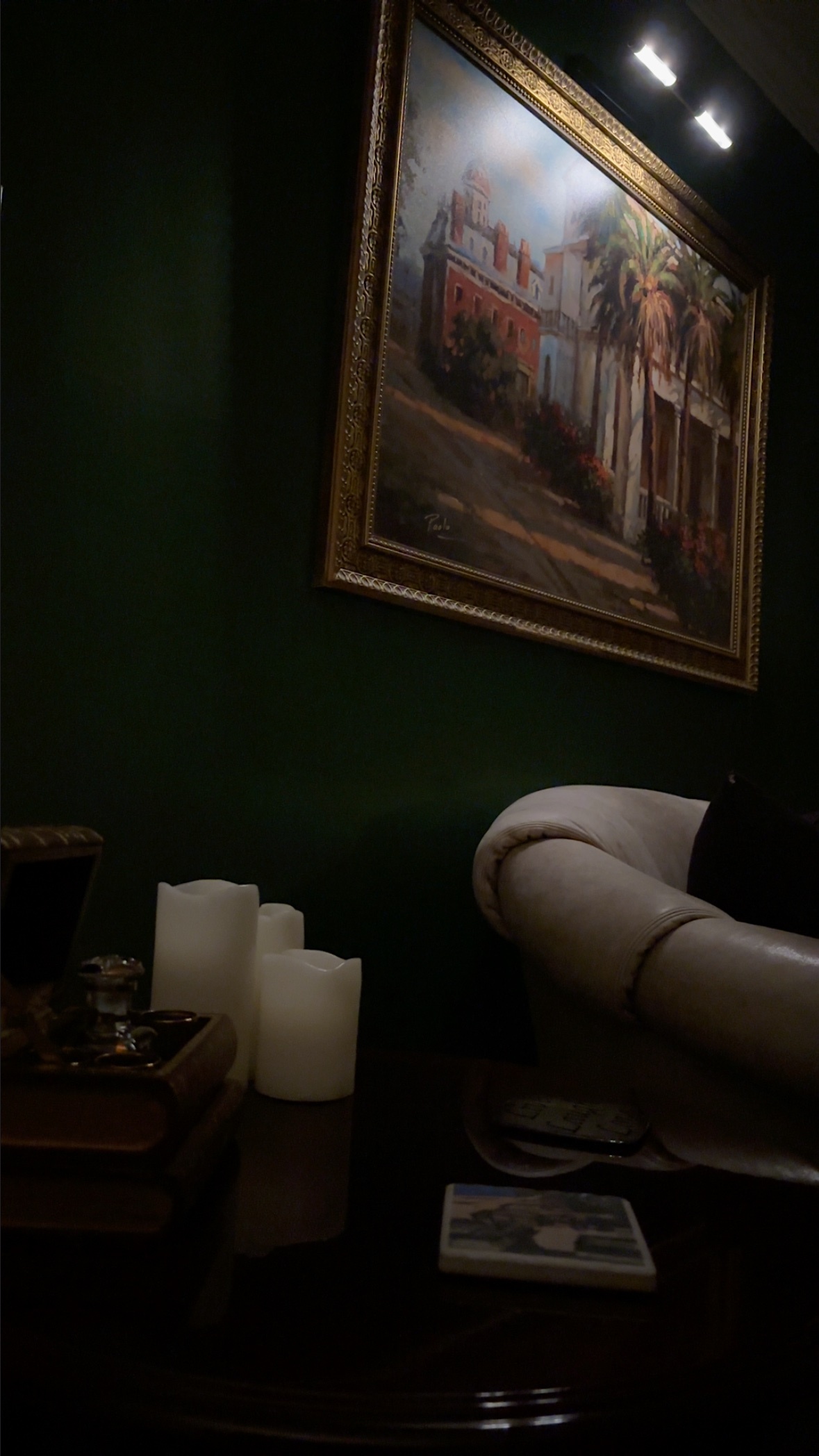 A painting of a building with palm trees hangs on a dark green wall above a table with candles and books. The room has a moody, vintage atmosphere.