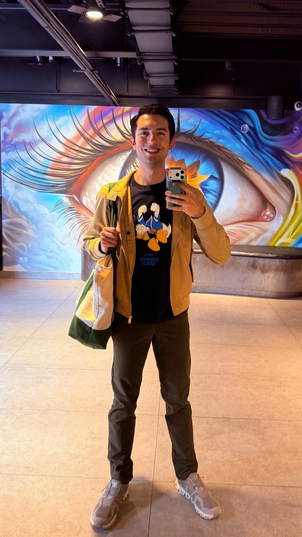 A man takes a selfie in front of a large, colorful mural of an eye. He is wearing a graphic t-shirt, a tan jacket, and dark pants, holding a tote bag.