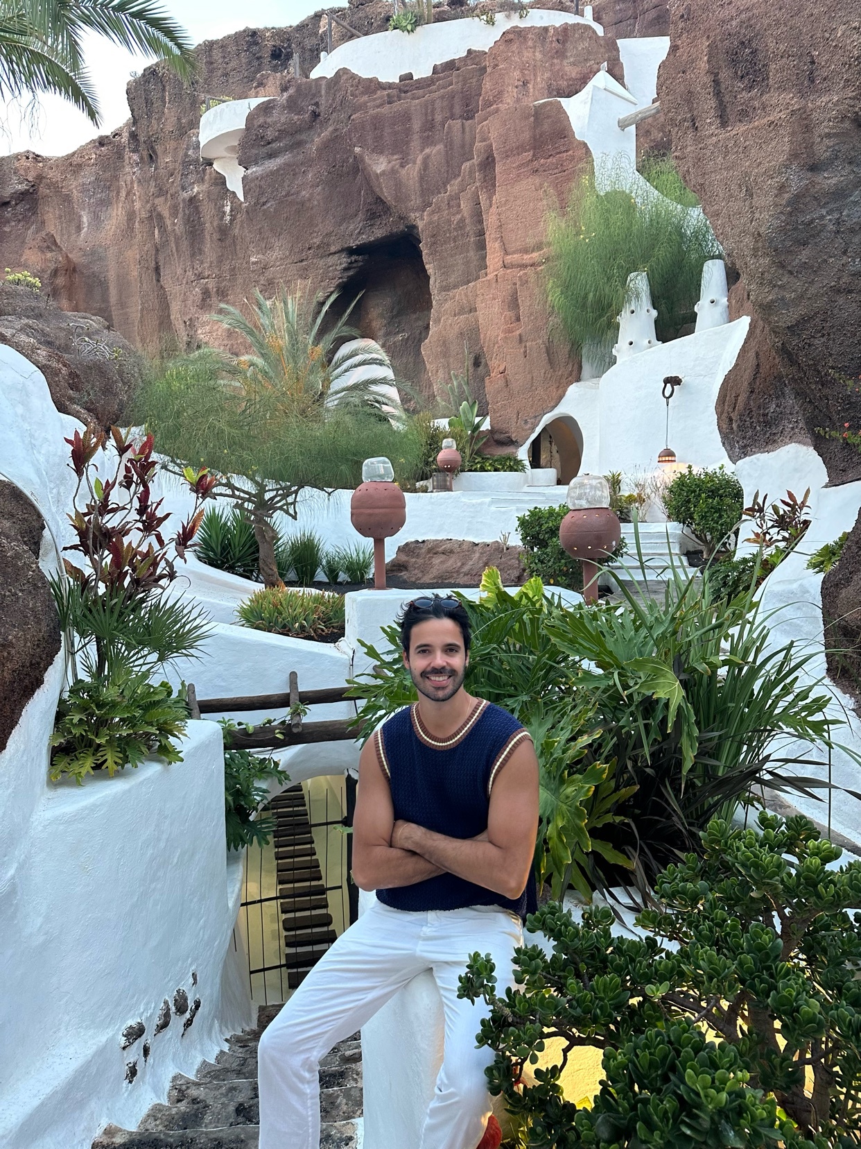 A man poses with his arms crossed in front of a unique white architectural structure built into a rocky hillside. Lush greenery surrounds the scene, suggesting a warm, vacation-like atmosphere.