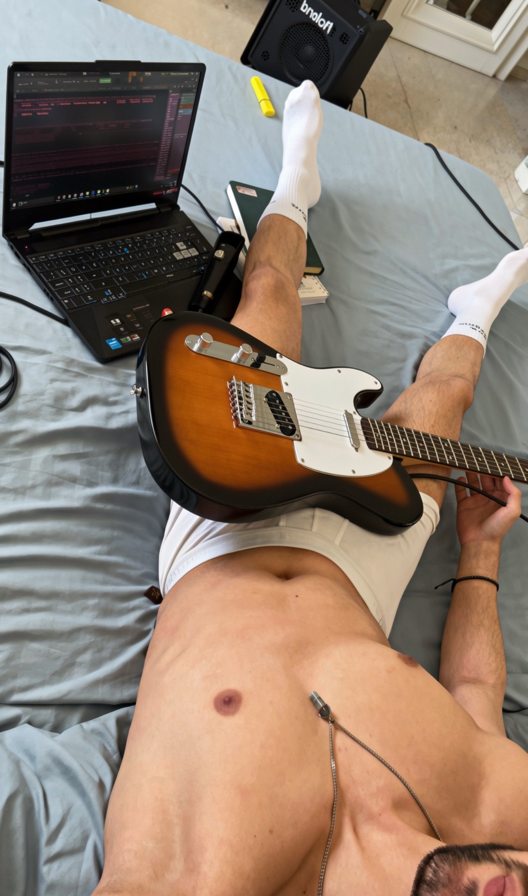 A shirtless man in underwear lies on a bed with a guitar across his lap. A laptop and other music equipment are nearby.