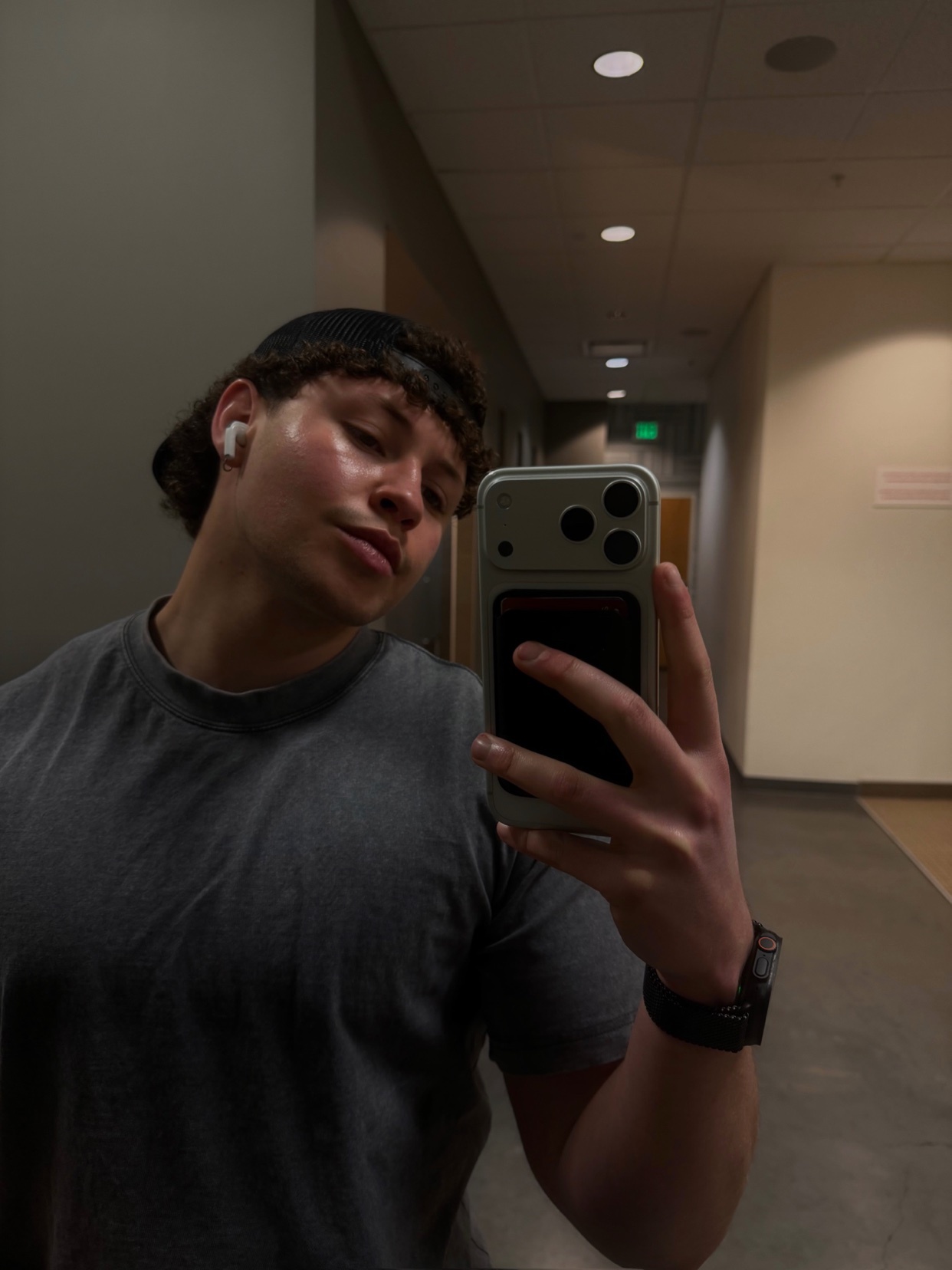 A man takes a selfie in a gym hallway, wearing a grey t-shirt, a hat, and earbuds. He appears ready for summer.