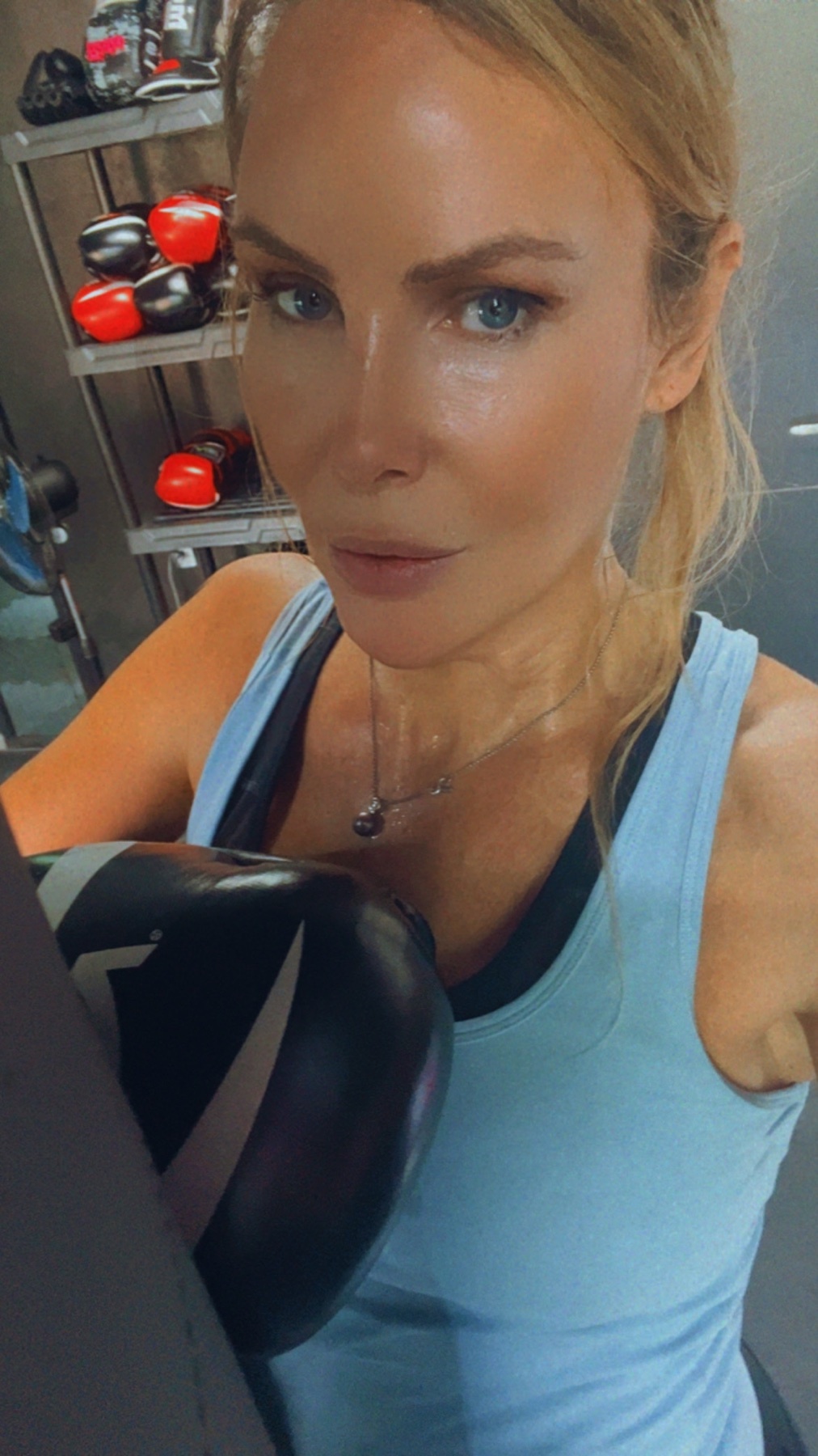 A woman with blonde hair and blue eyes poses for a selfie in a gym, wearing a light blue tank top and holding a boxing glove. She appears sweaty, indicating a recent workout.