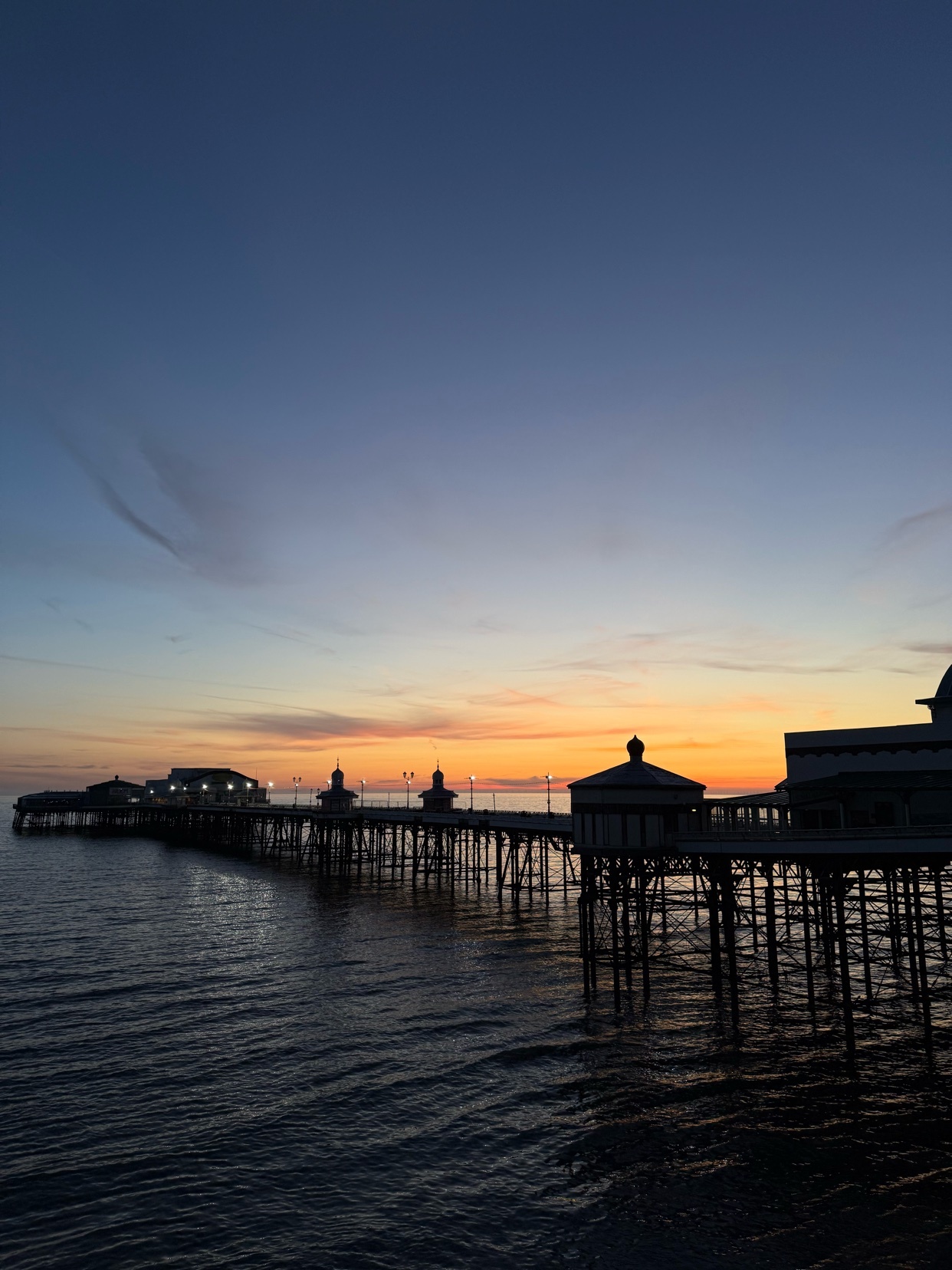 A pier extends into the ocean at dusk, silhouetted against a colorful sunset. The sky transitions from blue to orange, reflecting in the water.