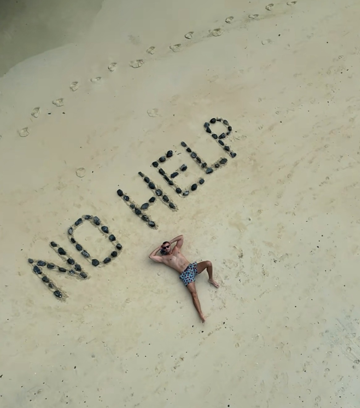 A man lies on the sand at the beach, with the words "NO HELP" written in sunglasses behind him. He is wearing patterned swim trunks and sunglasses, and appears to be relaxing.