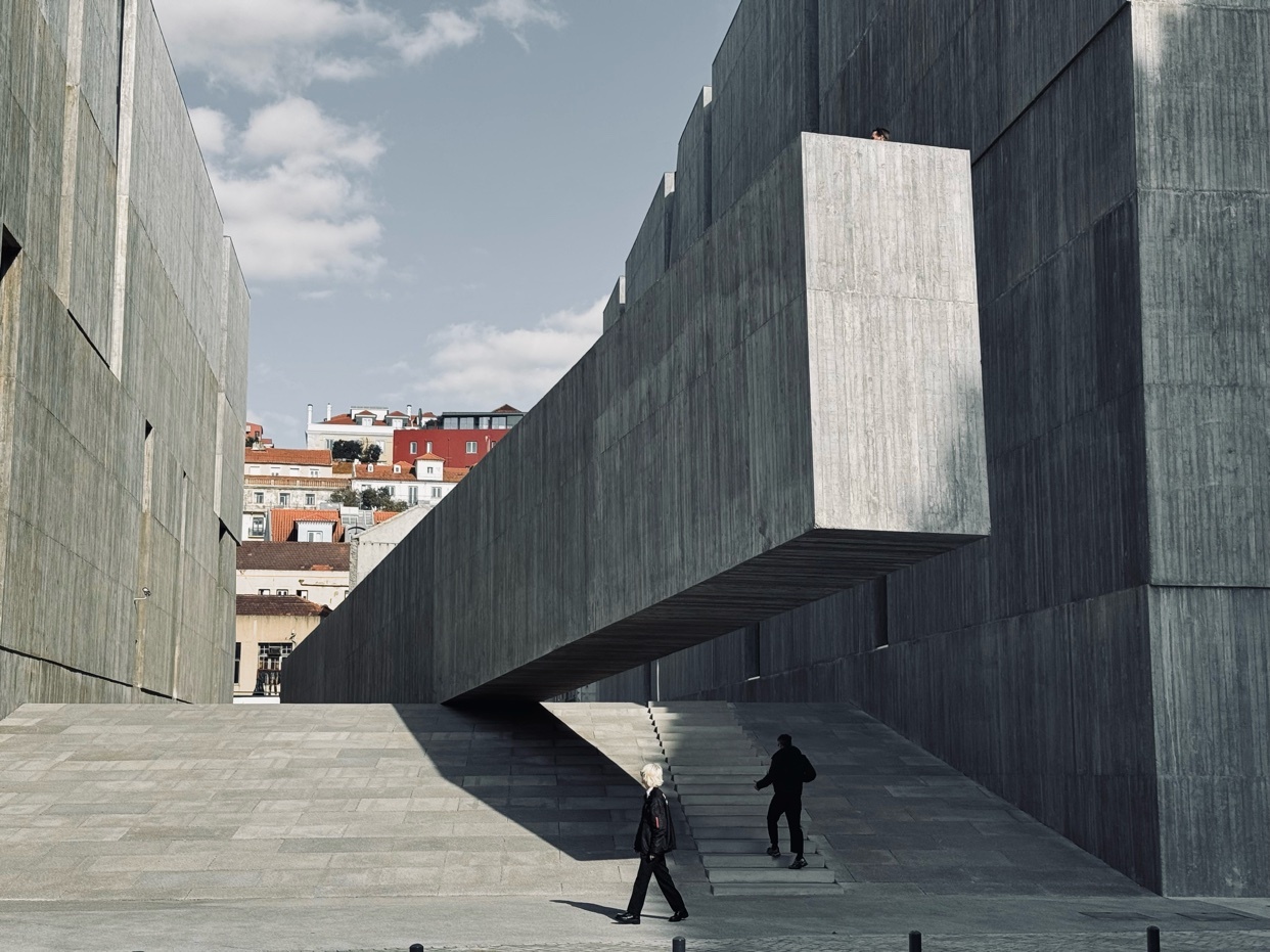 A striking architectural shot featuring a modern concrete building with a unique cantilevered design. Two people are walking in the foreground, adding scale and context to the urban scene.