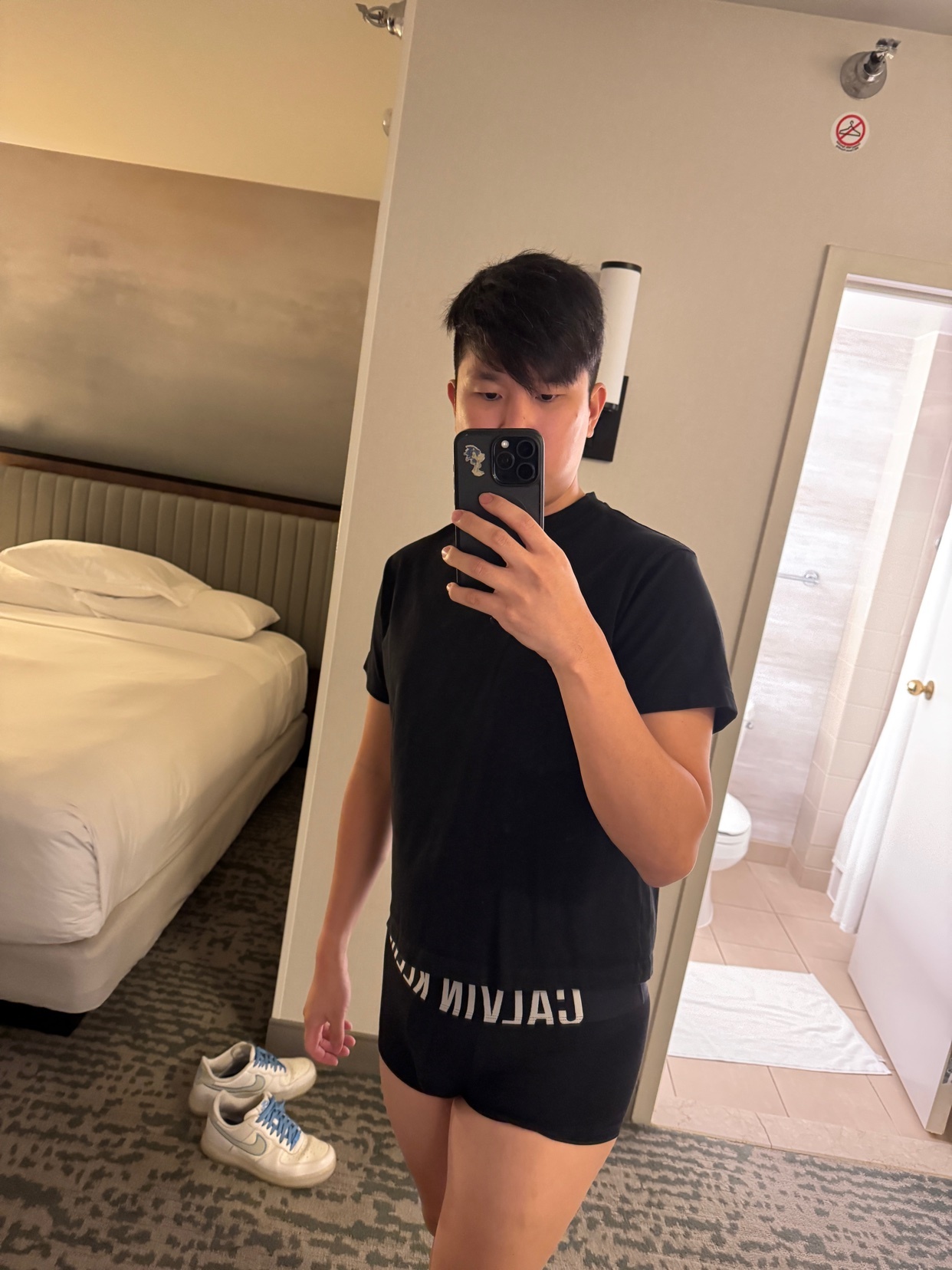 A man in a black t-shirt and Calvin Klein underwear takes a mirror selfie in a hotel room. His sneakers are on the floor next to the bed.