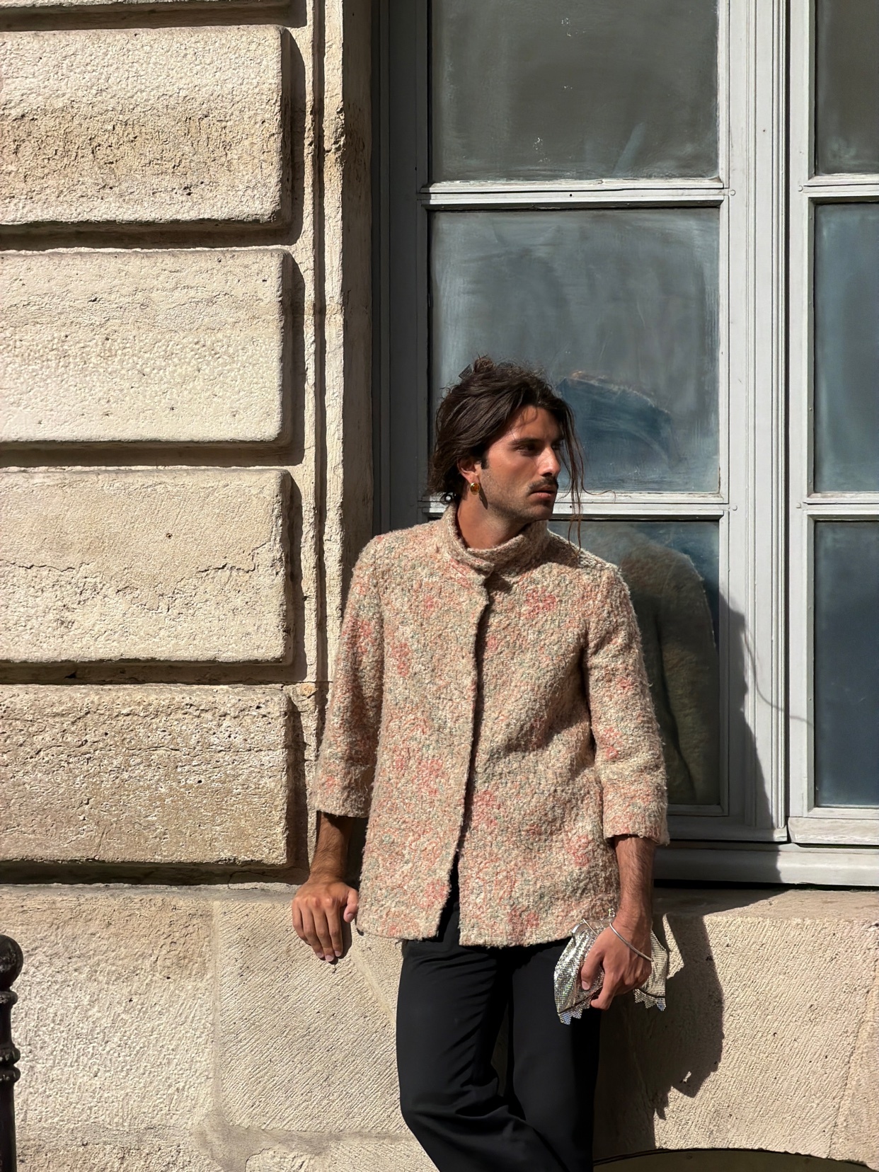 A man with long, dark hair leans against a stone building next to a large window. He is wearing a textured, patterned jacket and black pants, holding a small metallic accessory in his hand.