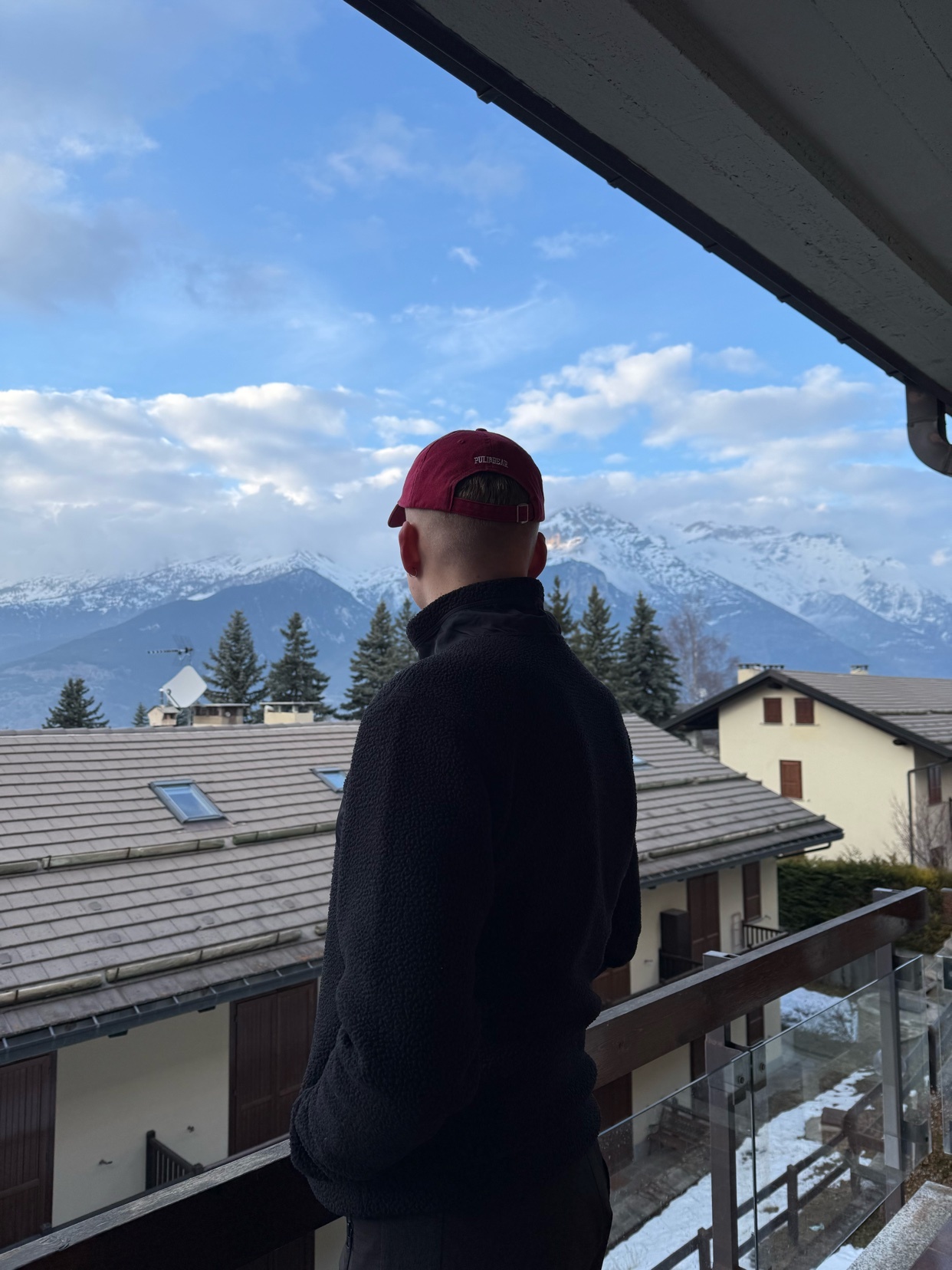 A man wearing a red baseball cap and a dark jacket stands on a balcony, looking out at a snowy mountain range under a cloudy sky.