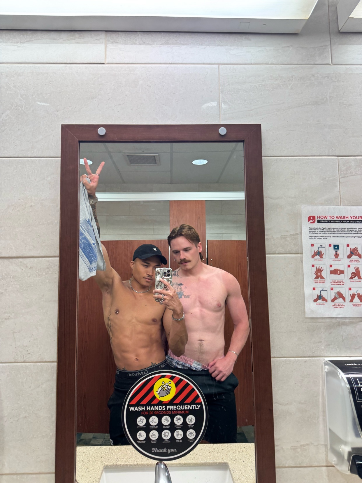 Two men pose for a mirror selfie in a restroom. One man on the left is wearing a black baseball cap and holding up a bag, while the man on the right has a mustache and is looking at the camera.
