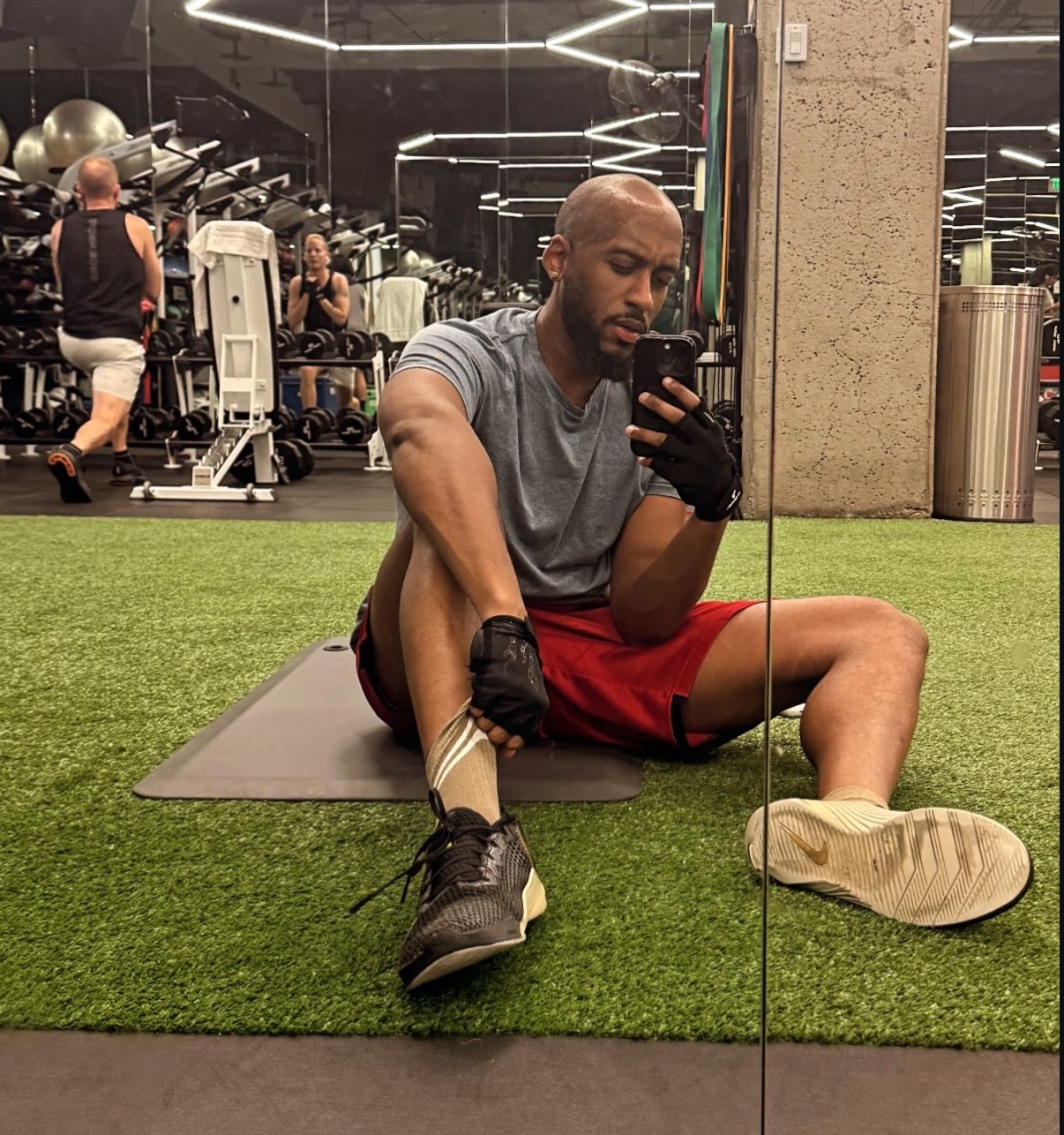 A man takes a mirror selfie at the gym. He is shirtless and wearing shorts.