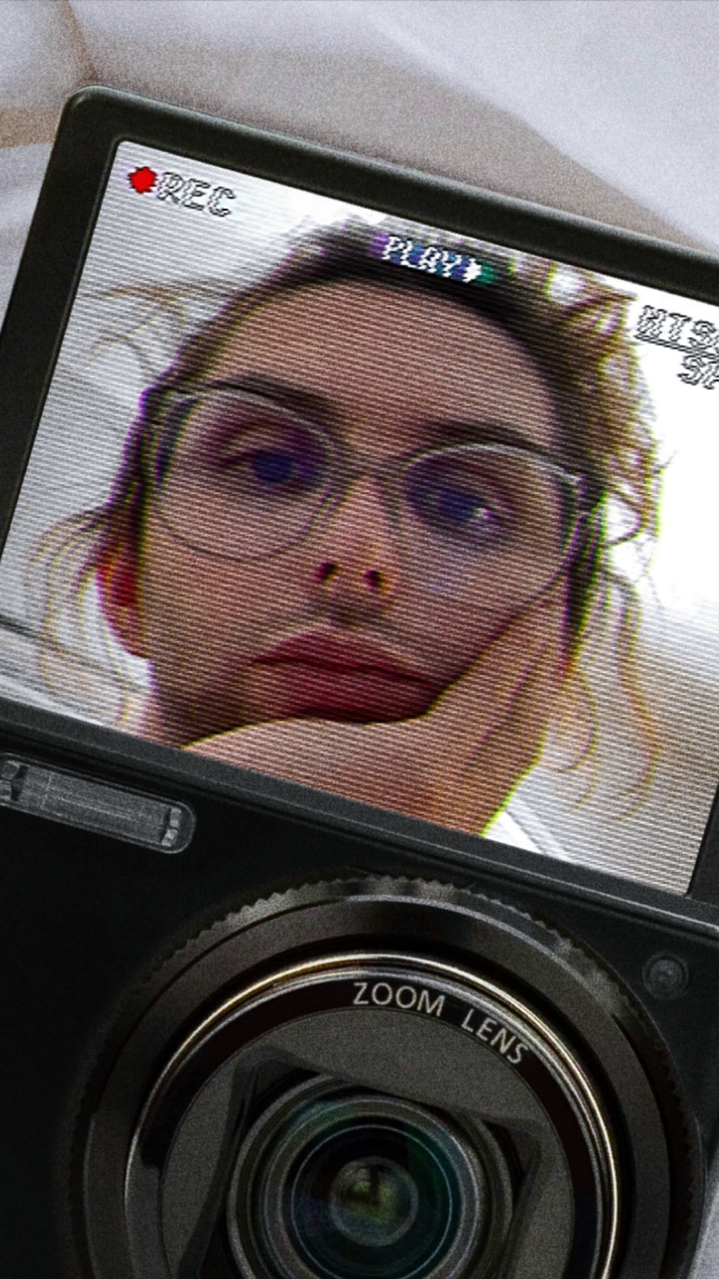 A man with long hair and glasses is taking a selfie with a vlog camera. The camera screen shows a recording indicator and a play button.