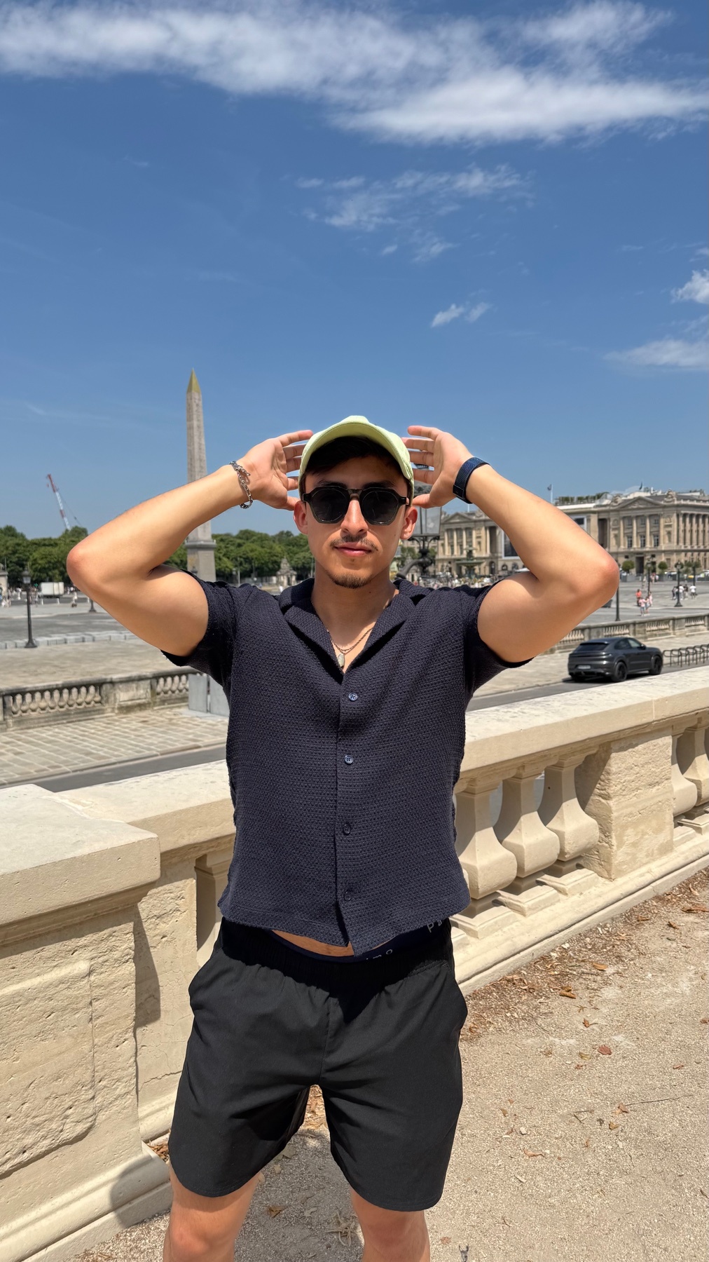 A man poses for a photo outdoors in front of a cityscape, wearing sunglasses and a hat.