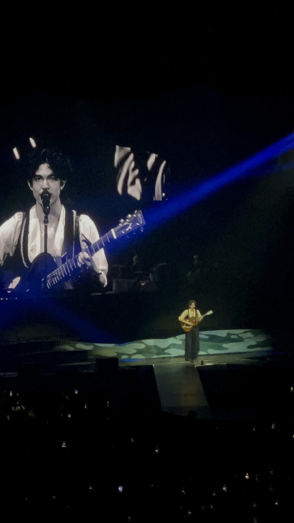 A man plays guitar and sings on a brightly lit stage, with a large screen behind him displaying a close-up of his face. The audience is visible in the foreground, with small lights twinkling in the darkness.