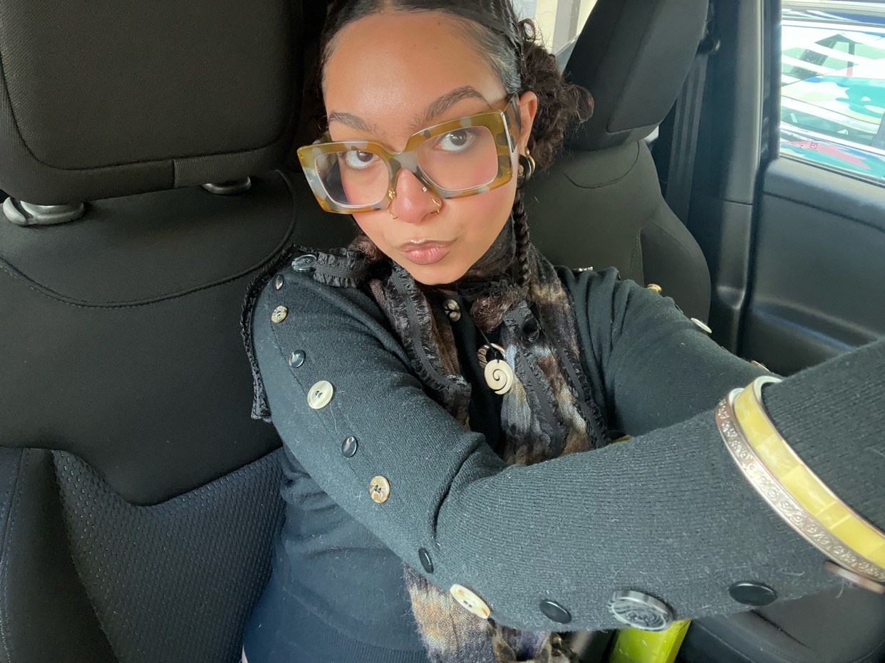 A woman takes a selfie while wearing large glasses and a stylish outfit. She is sitting in a car.