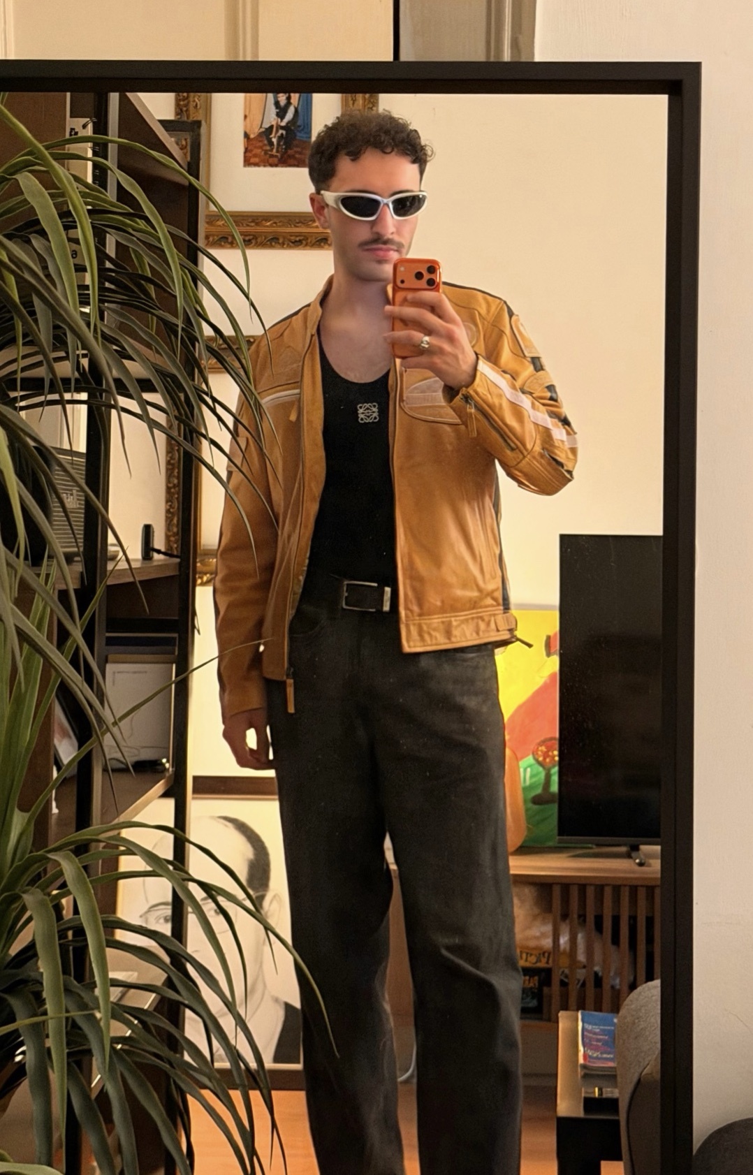A man in a yellow leather jacket and sunglasses takes a mirror selfie.