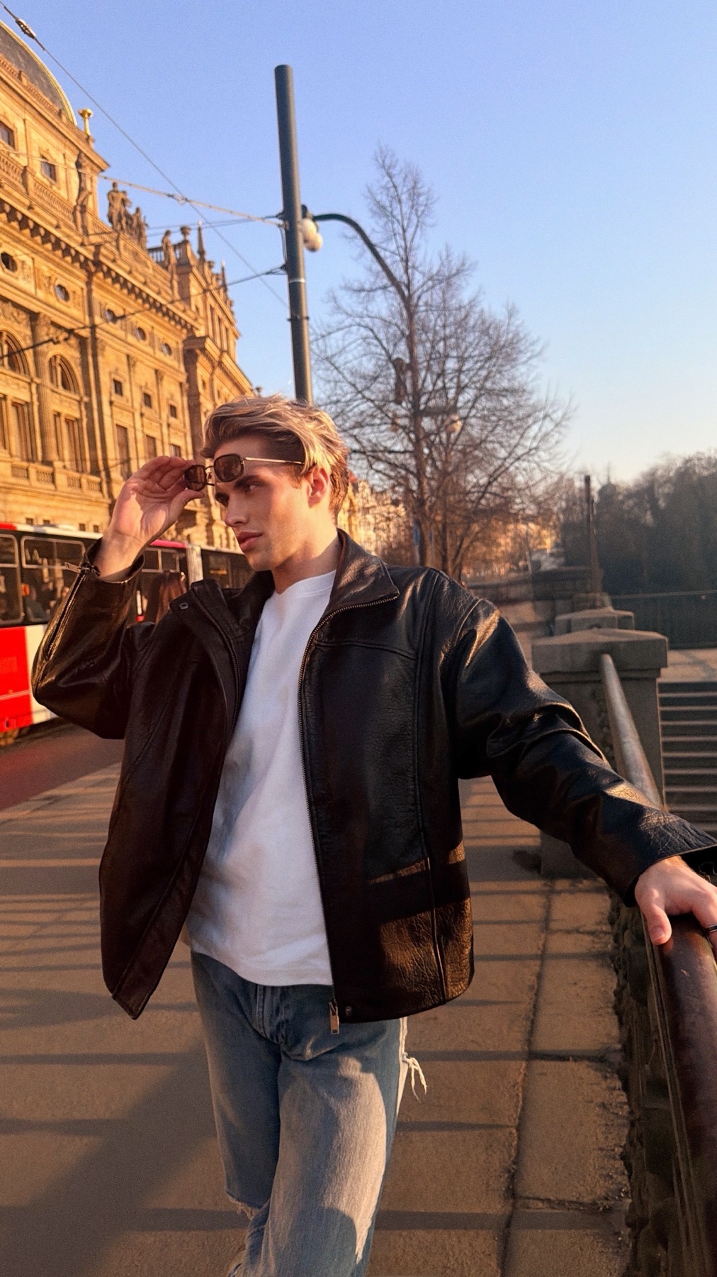 A young man in sunglasses and a leather jacket poses on a city street with historic architecture in the background.