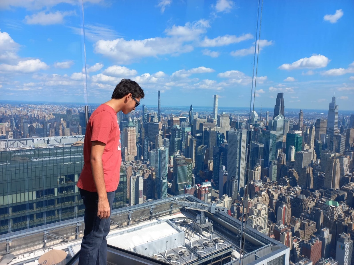 A man in a red t-shirt and jeans stands on a high-rise balcony overlooking the New York City skyline under a bright, cloudy sky.
