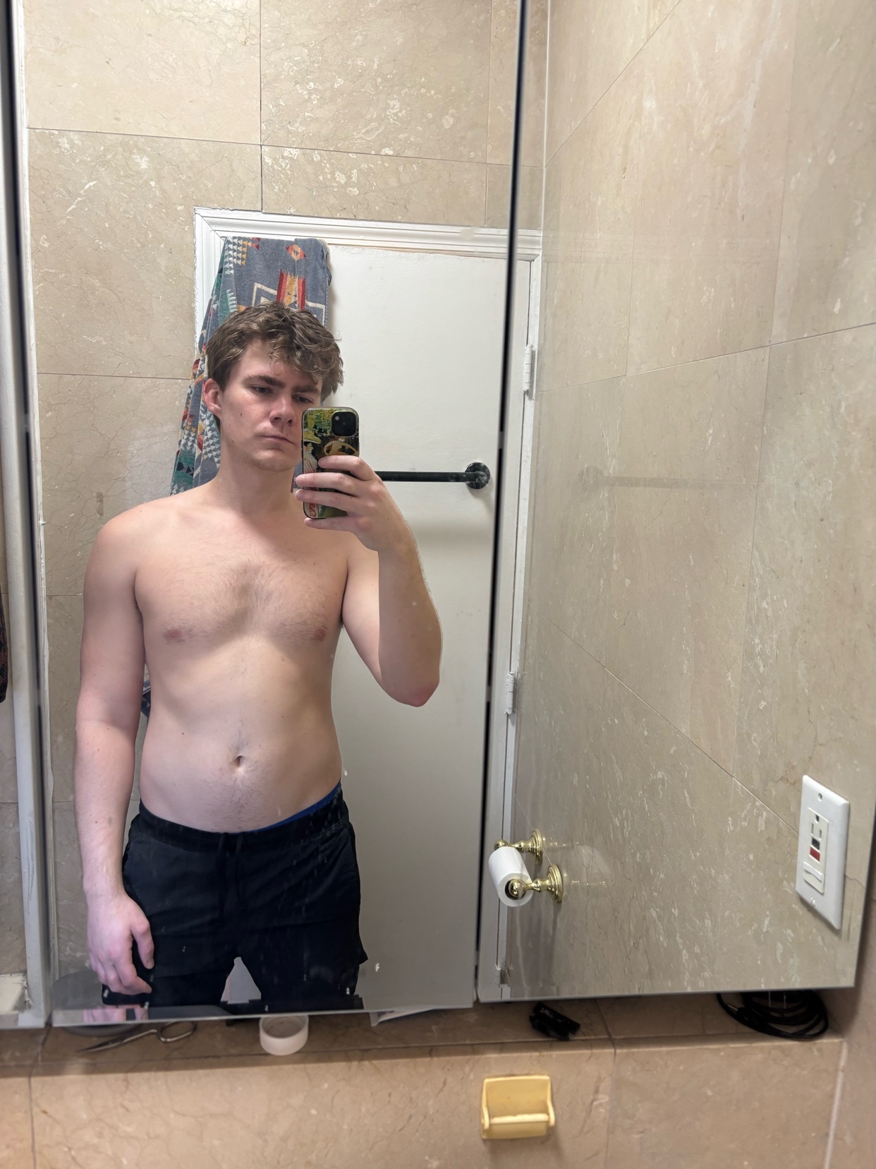 A young man takes a mirror selfie in a bathroom, wearing only shorts. He has light brown, messy hair and is looking at the camera.