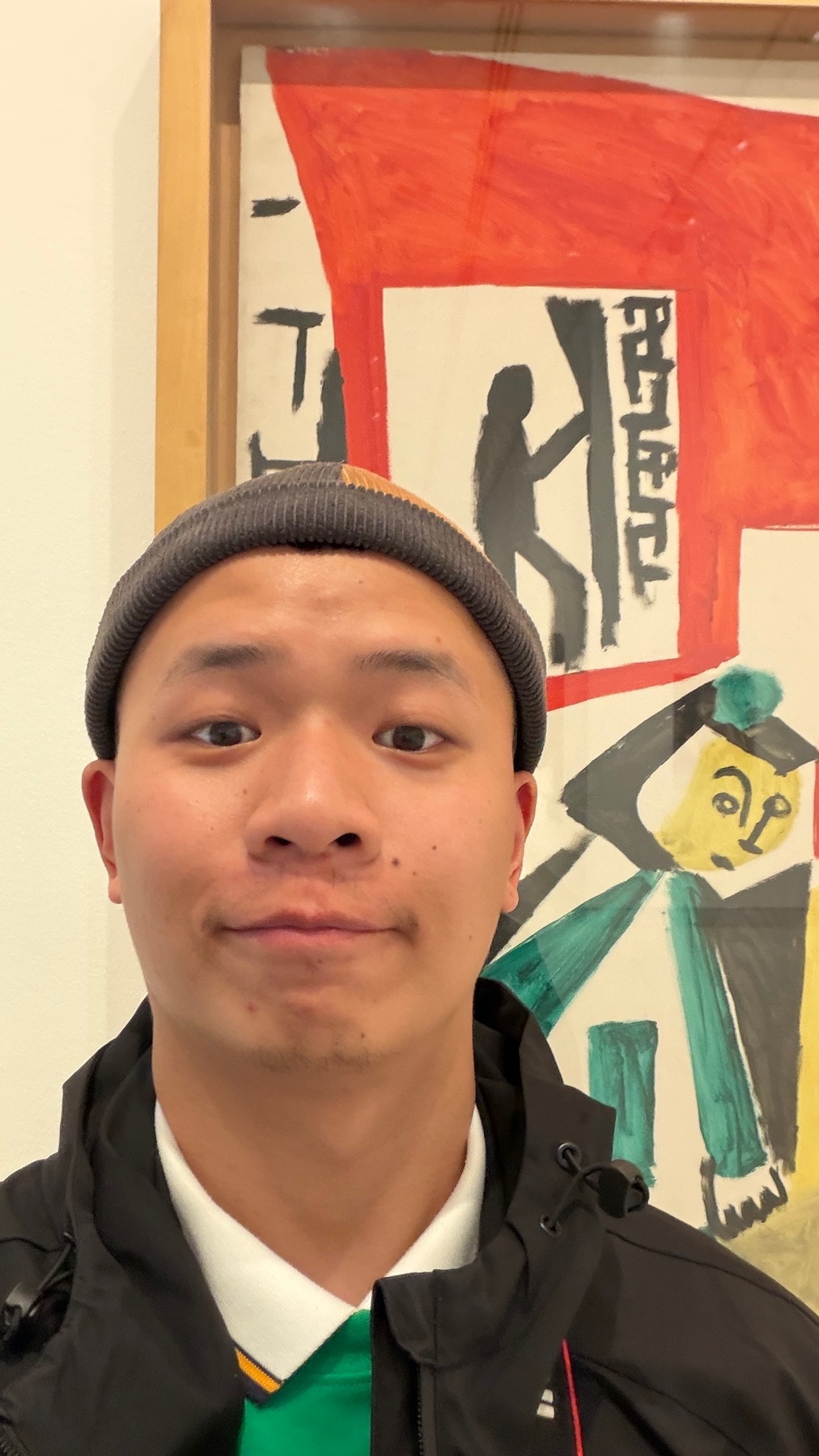 A man poses in front of a colorful painting at the Museo de Picasso. The man is wearing a beanie and a jacket.